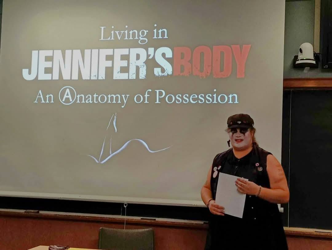 Person standing in front of a presentation slide, holding papers, wearing sunglasses and a black outfit.