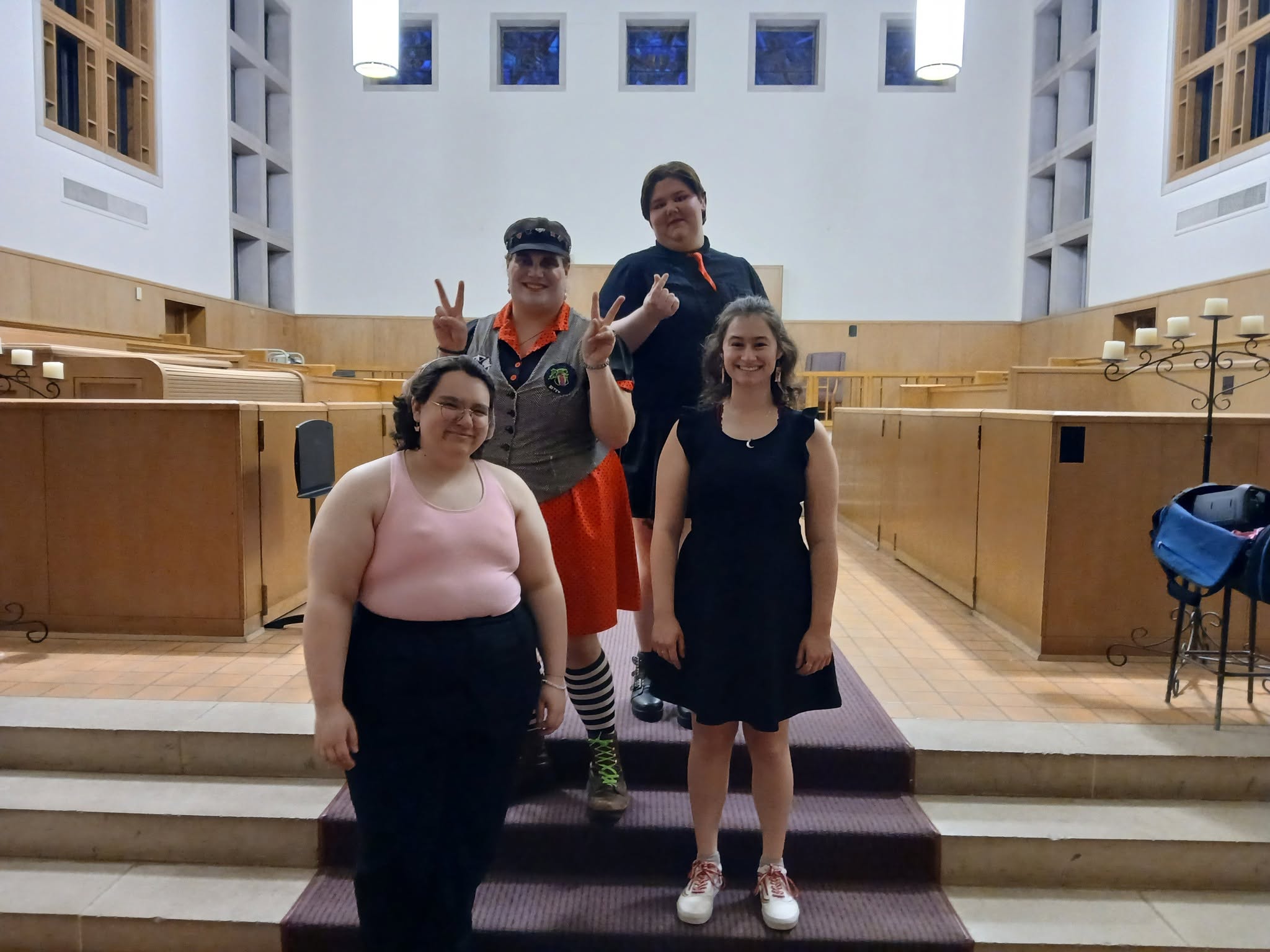 Four people standing on steps inside a bright room with wooden benches and white walls, some making peace signs.