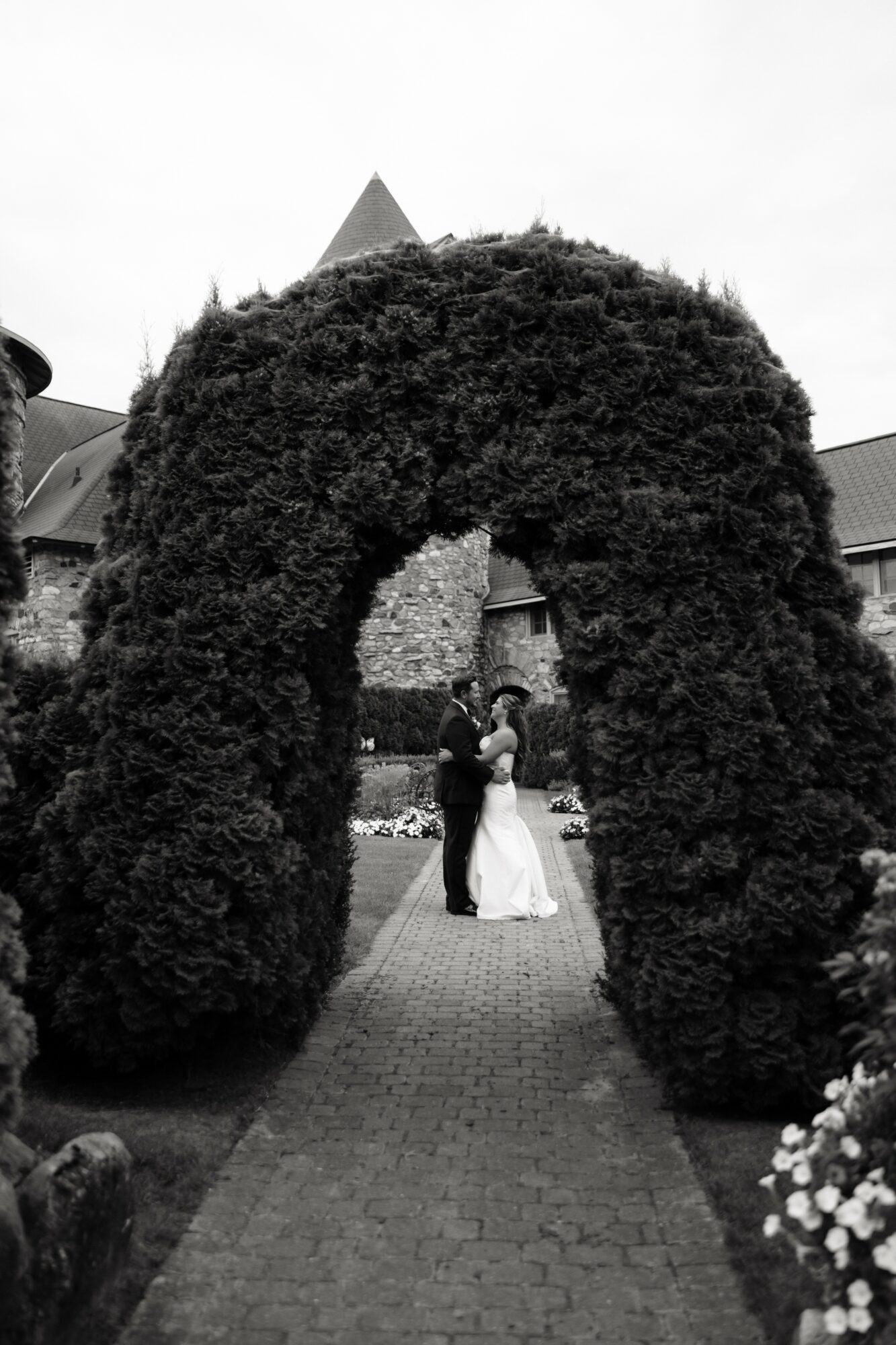 Couple dancing under a large, arched hedge in a garden, with buildings and a cloudy sky in background.
