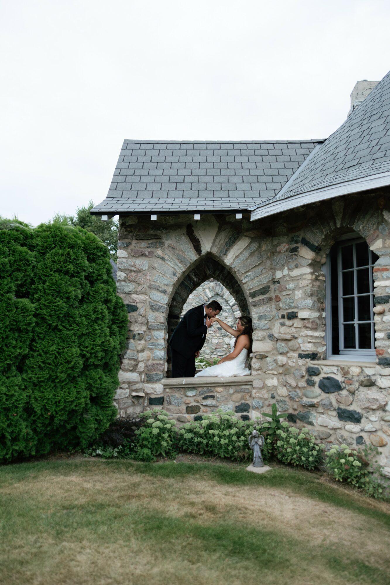 Two people stand in an arched window opening of a stone building, holding hands and smiling, with greenery in front.