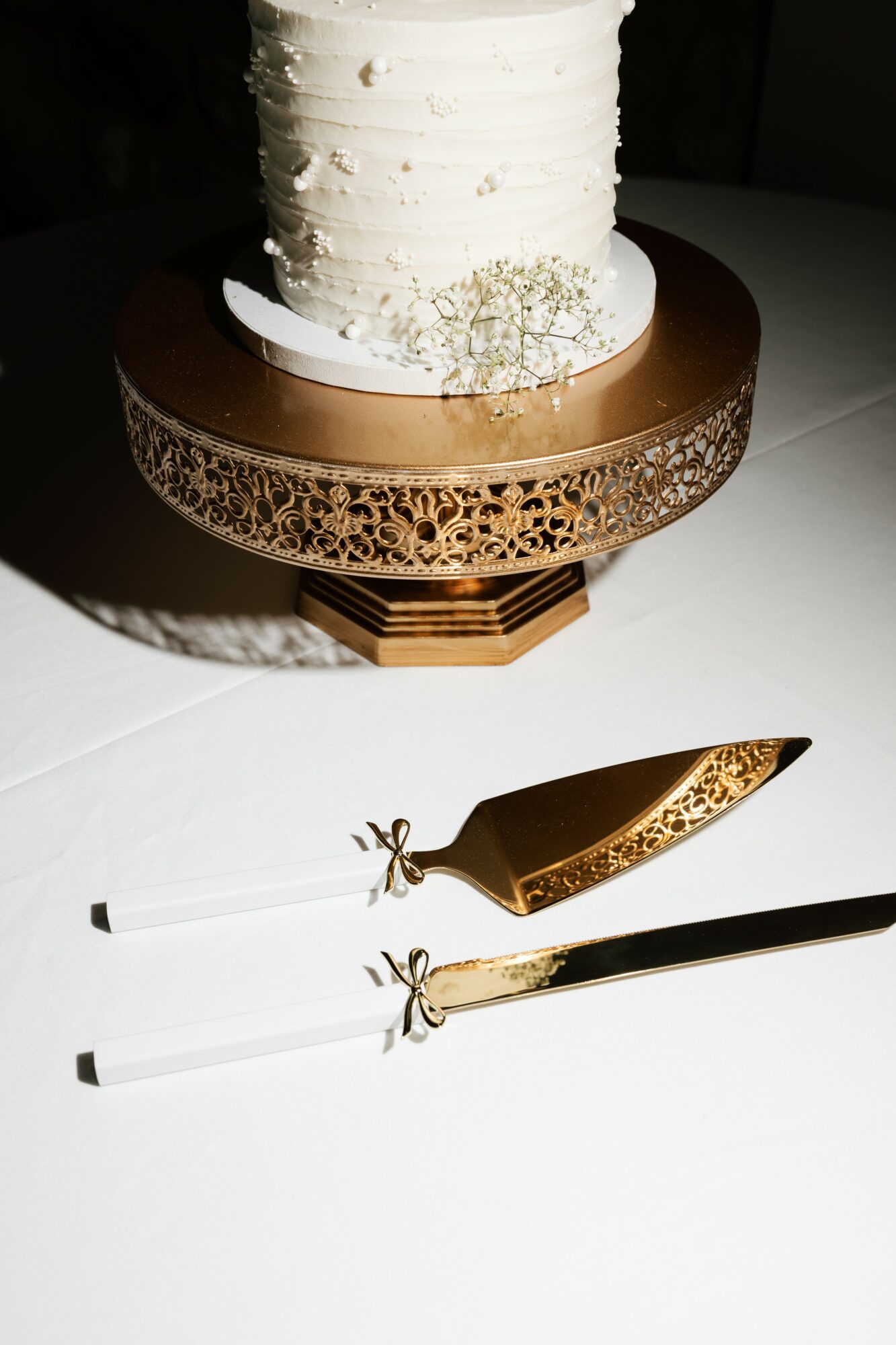 A white frosted cake on a decorative gold and brown stand with a gold knife and two white utensils beside it.