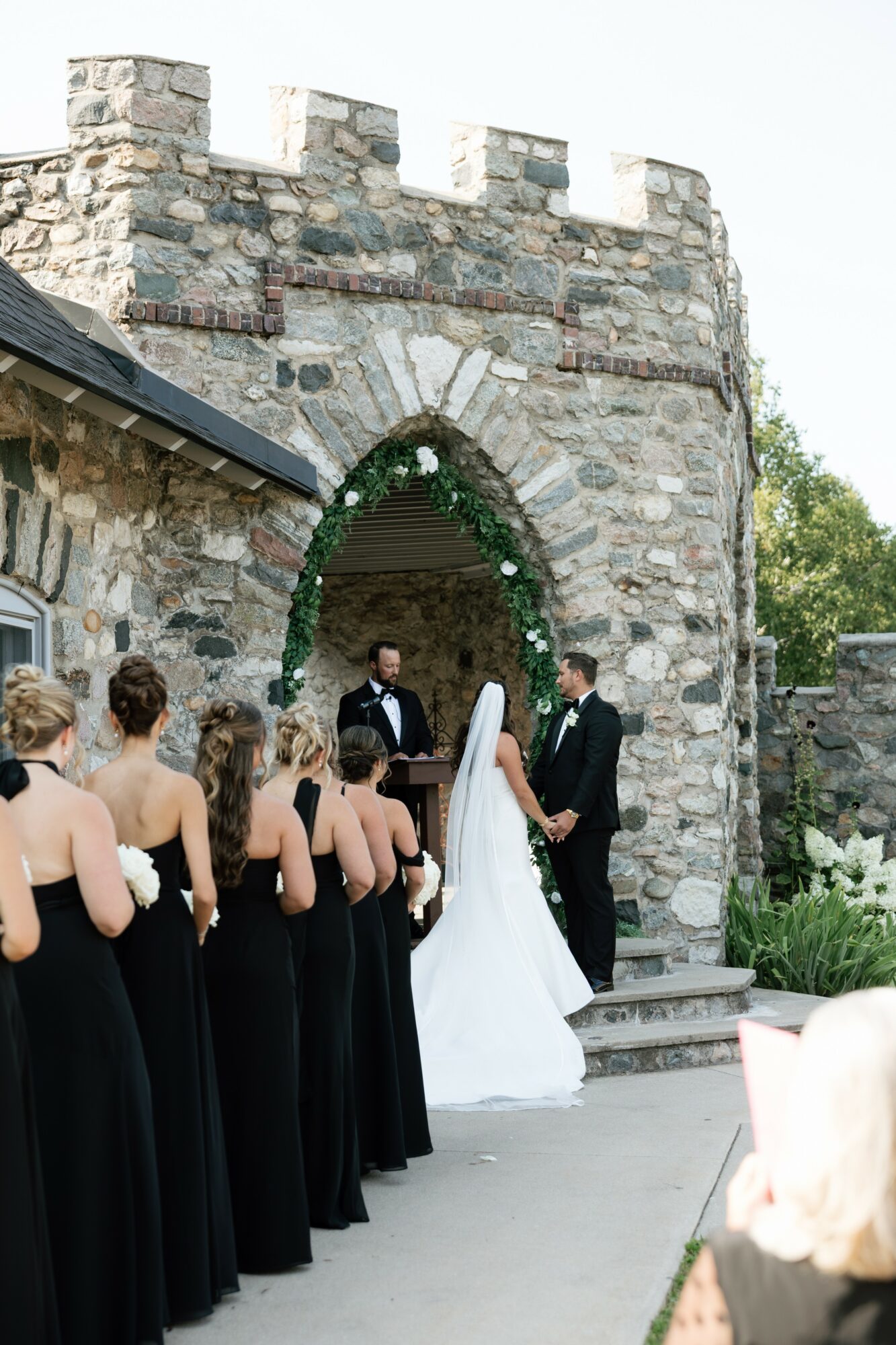 Bride and groom stand under an arched stone doorway during wedding ceremony, guests in black dresses watch, outdoor setting.