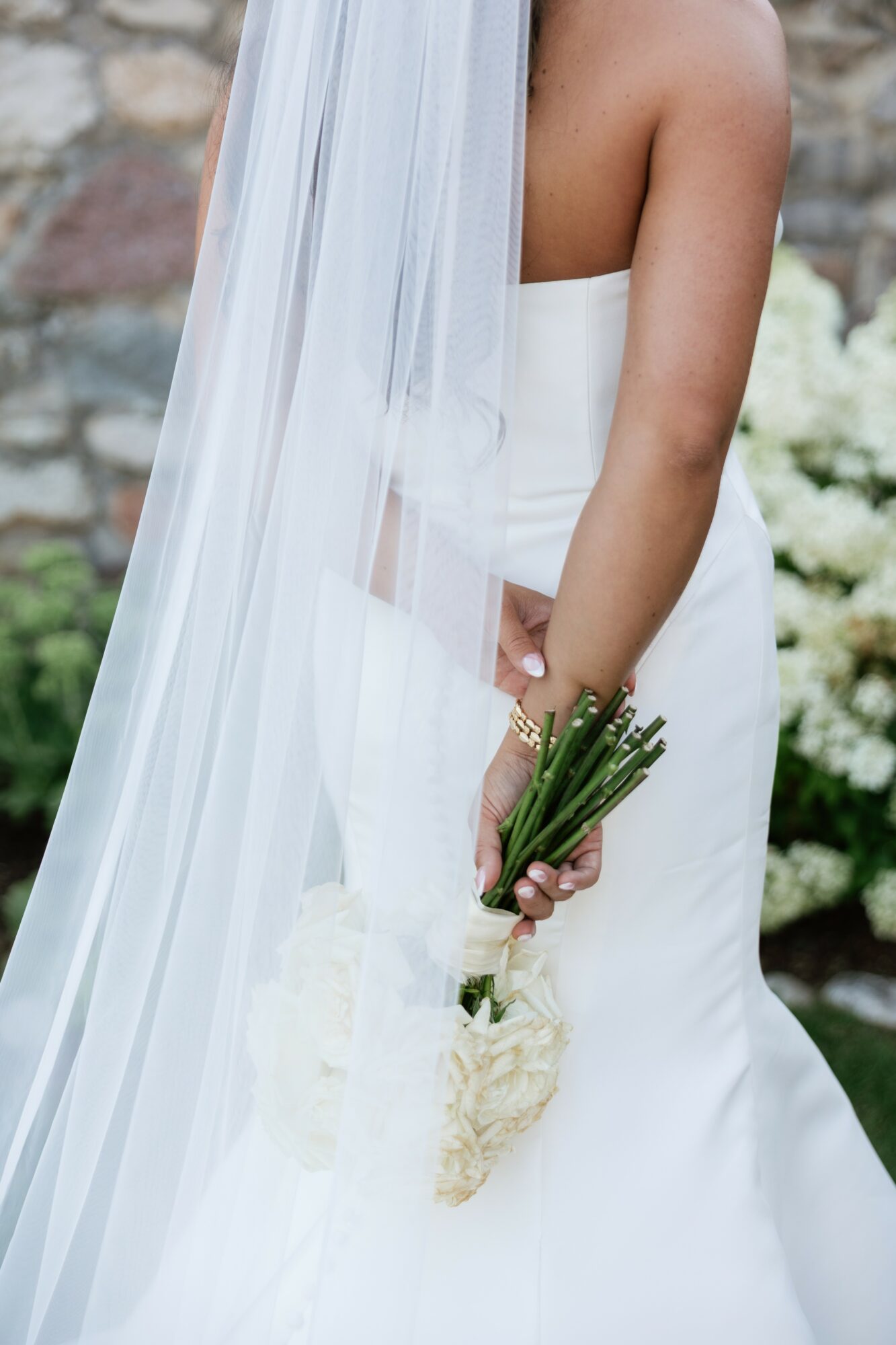 Close-up of a bride holding a bouquet of white flowers, wearing a strapless wedding dress and veil, outdoors with greenery.