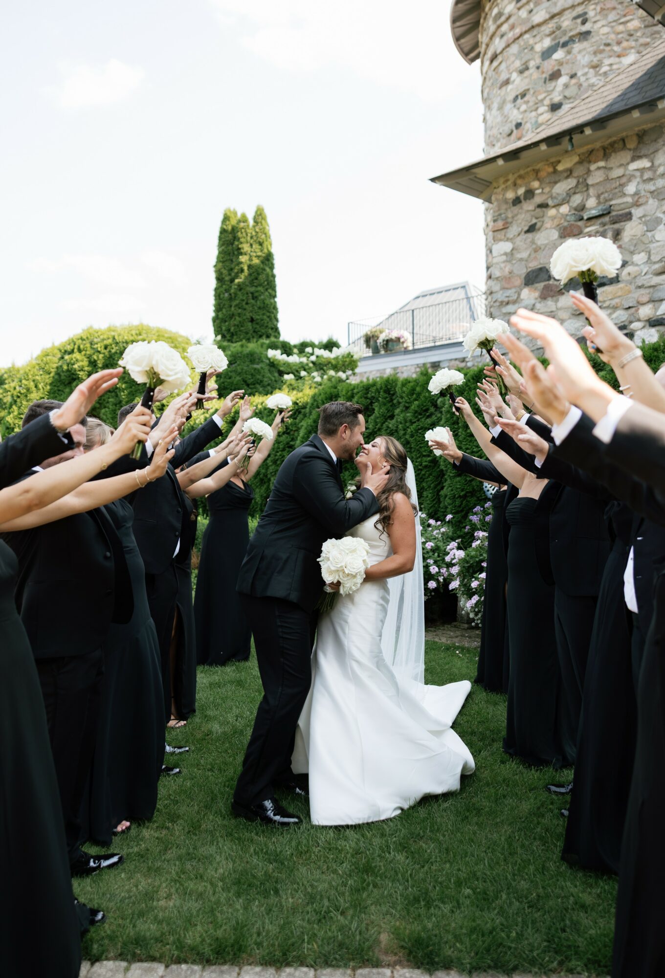 Bride and groom kissing outdoors, surrounded by people holding bouquets, near a stone building and greenery.