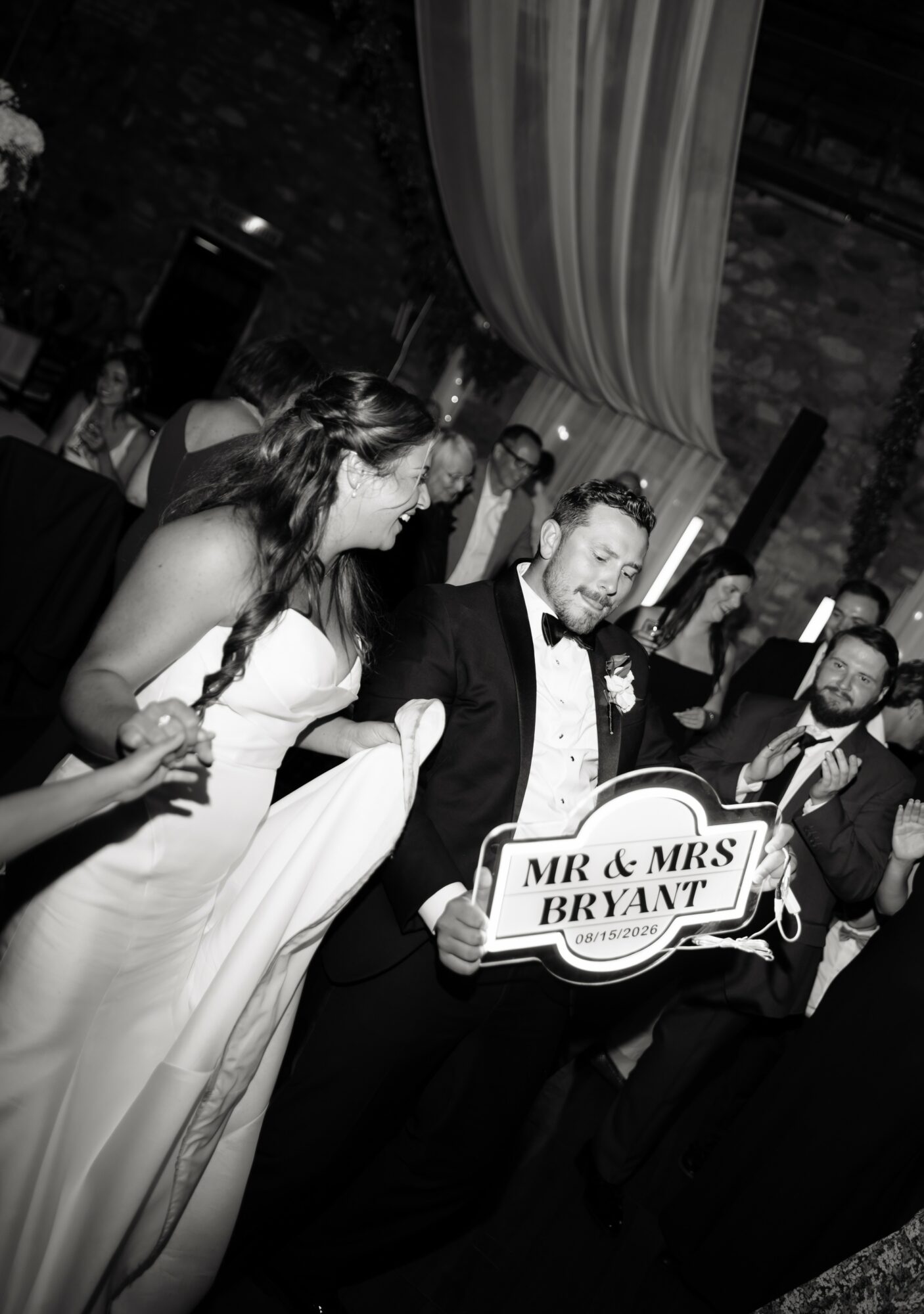 Group of people at a wedding reception, with a man holding a sign that says 'MR & MRS BRYANT', in a decorated venue.