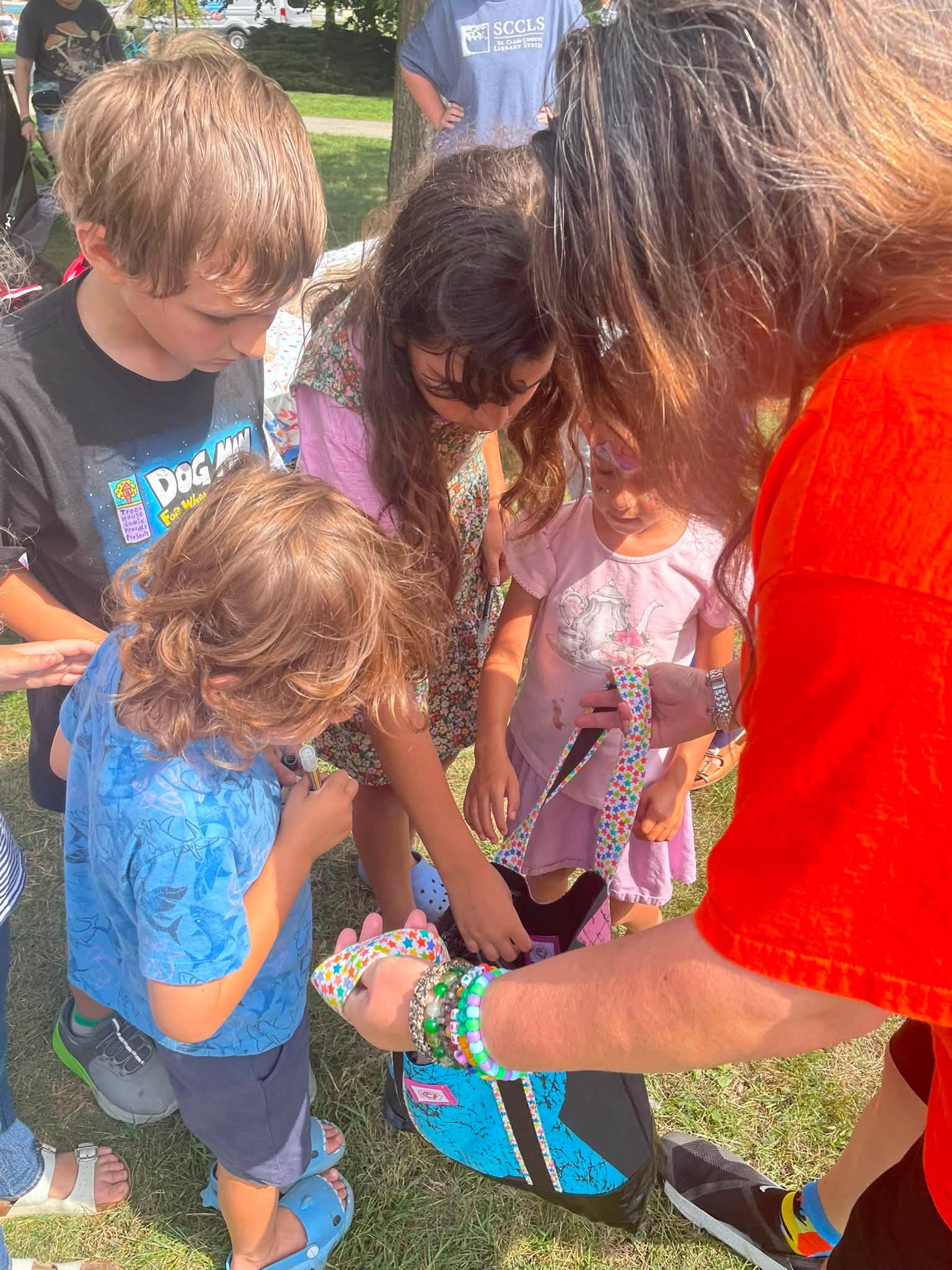 Group of children and an adult gathered outdoors around a backpack, engaging with its contents, on grass with trees in background.