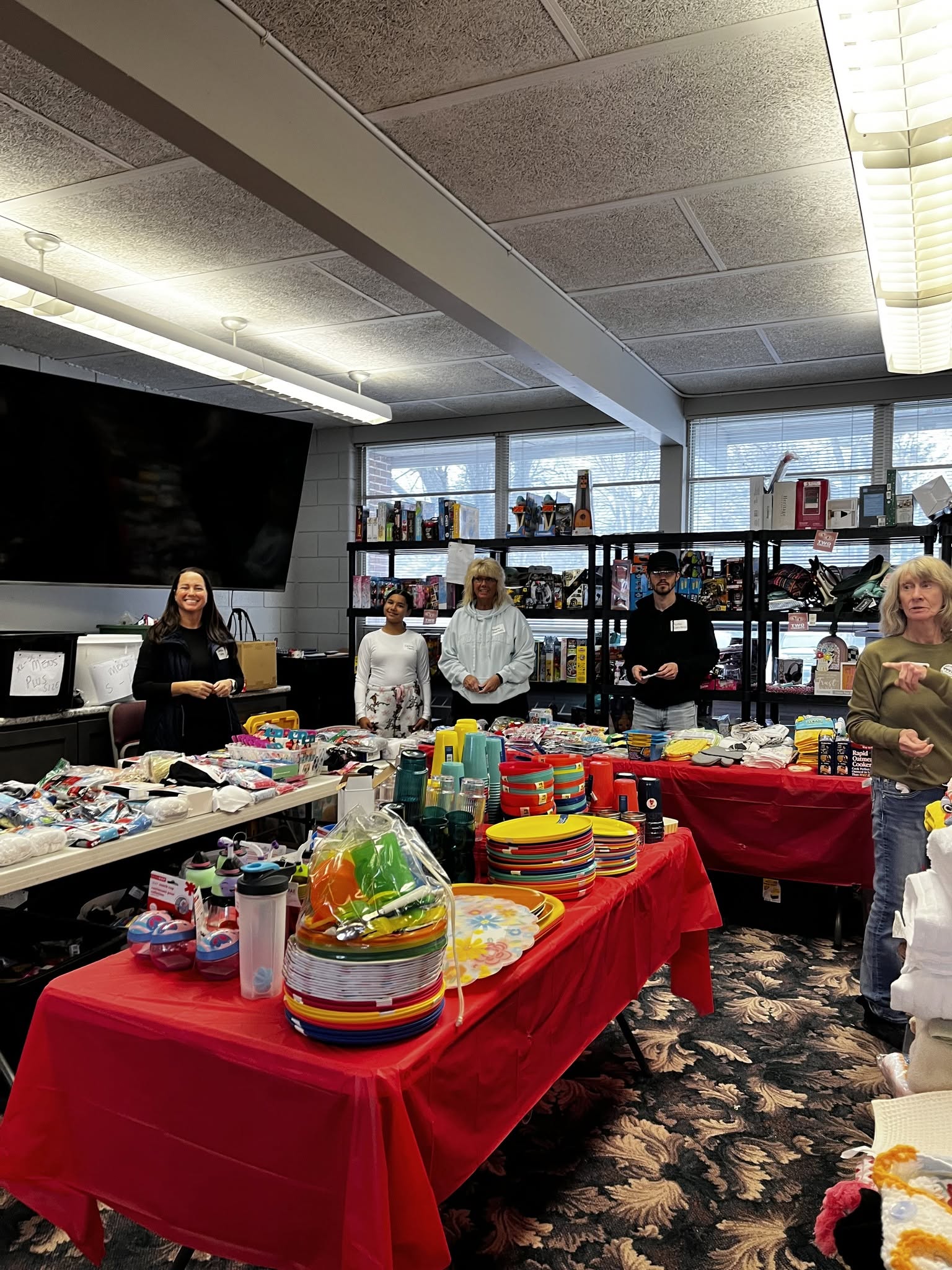 People standing behind tables with various items in a room with large windows and shelves, red tablecloths, and a patterned carpet.