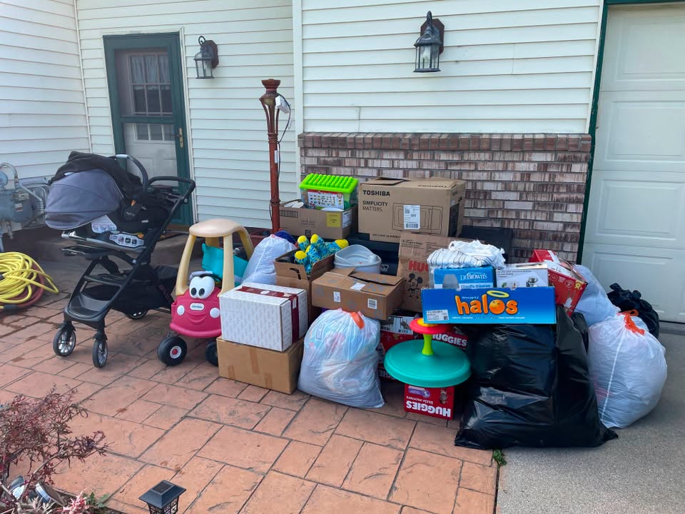 Cluttered driveway with boxes, bags, and children's toys in front of house with light blue siding and brick accents.