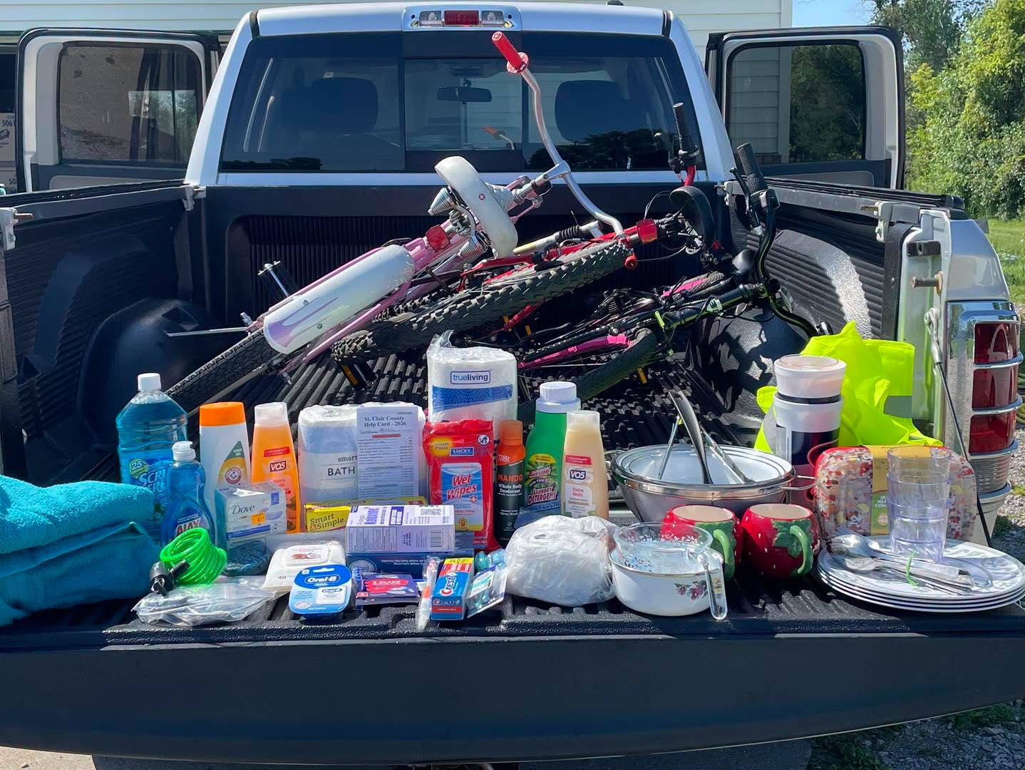 Pickup truck with bicycle and various bottles, containers, and supplies in the truck bed, outdoors on a sunny day.