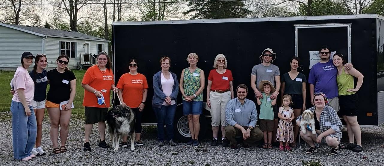 Group of people standing outdoors in front of a large black truck, some holding dogs, with trees and buildings in background.