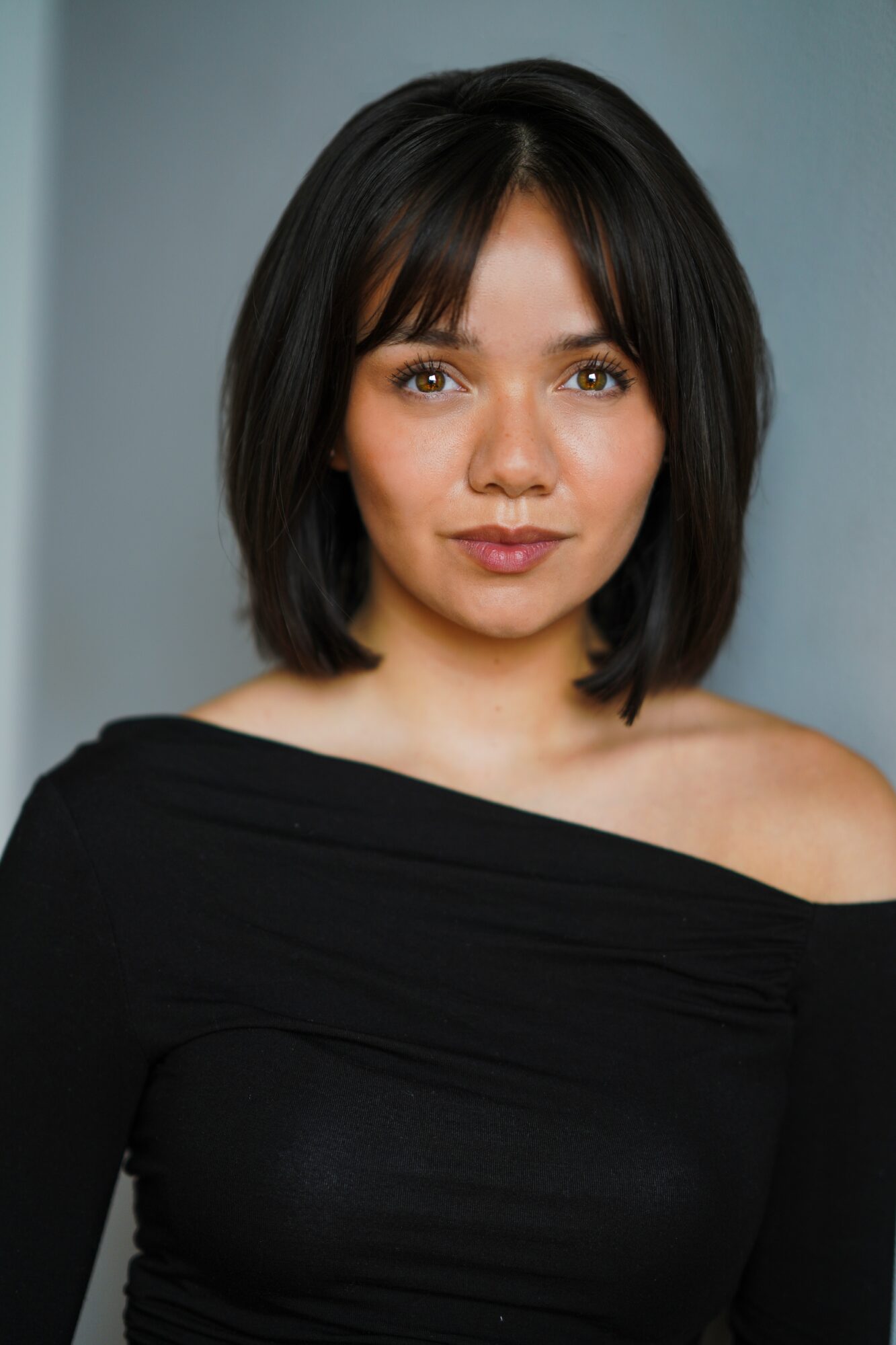 Young woman with shoulder-length dark hair and a black off-shoulder top, looking at the camera with a neutral expression.