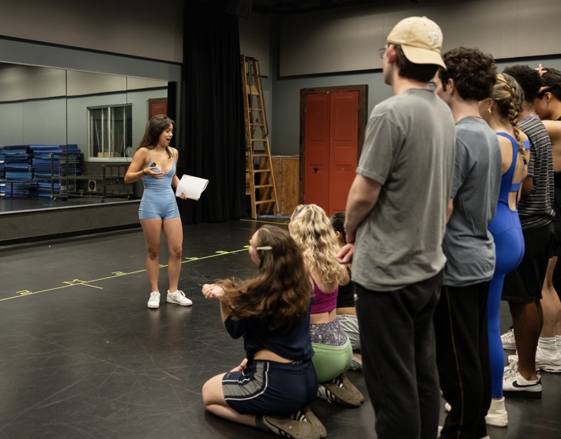 Group of people listening to a woman holding a paper in a rehearsal studio with mirrors and a ladder.