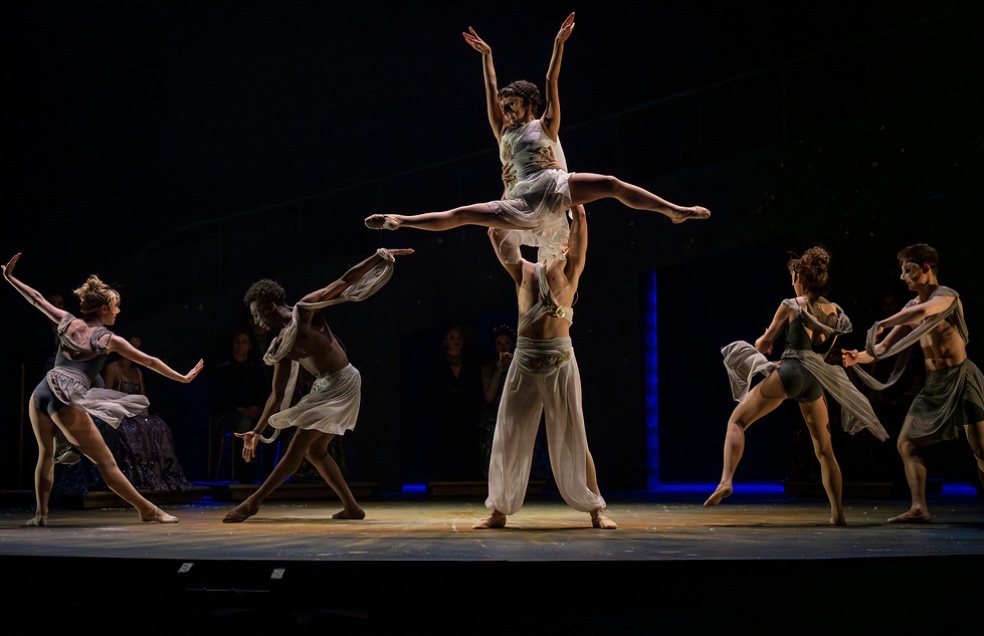 Ballet dancers perform on stage with one dancer lifted overhead, others in various poses, dark background, stage lighting.