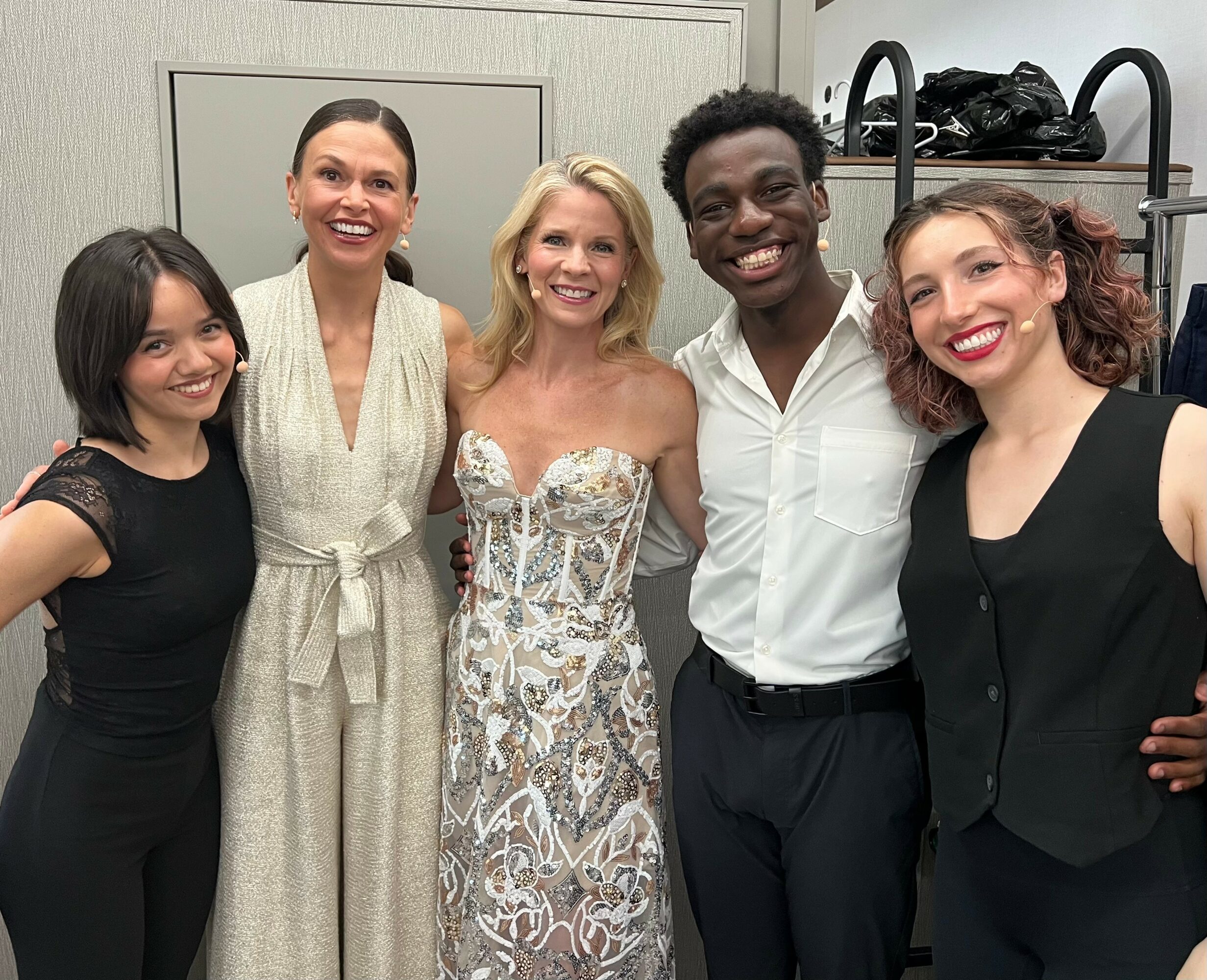 Five people standing together, smiling, in a room with a gray wall and shelves in the background.