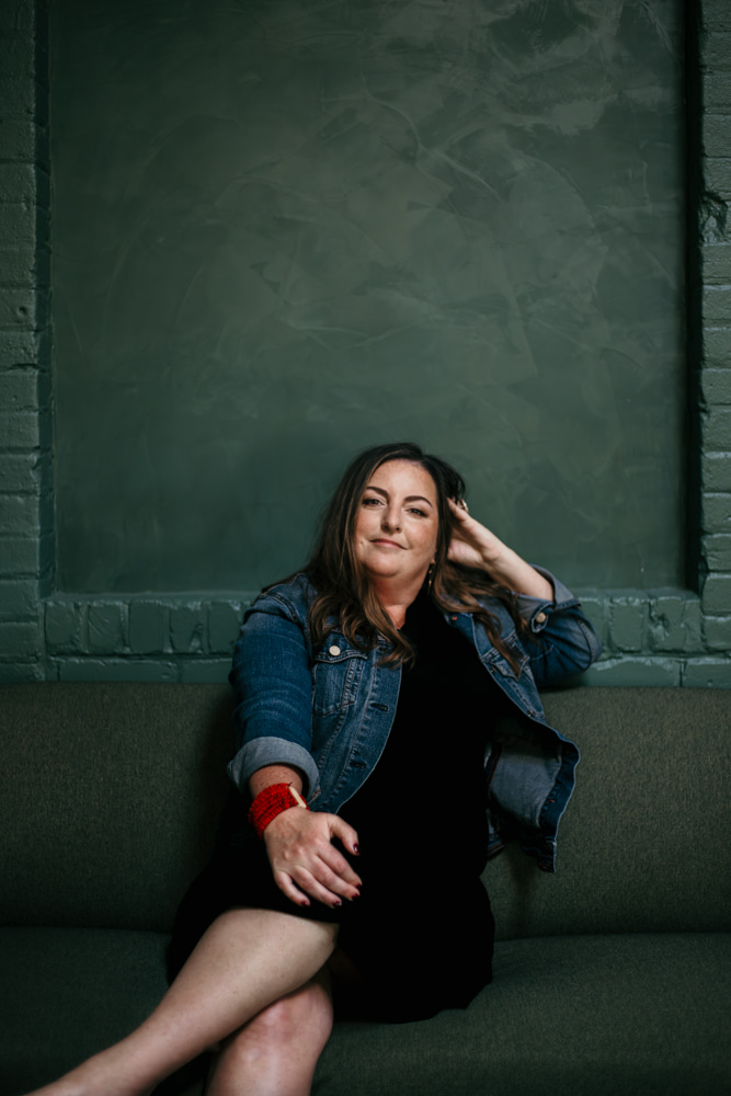 Woman sitting on a bench with a green wall background, wearing a denim jacket and black dress, smiling.