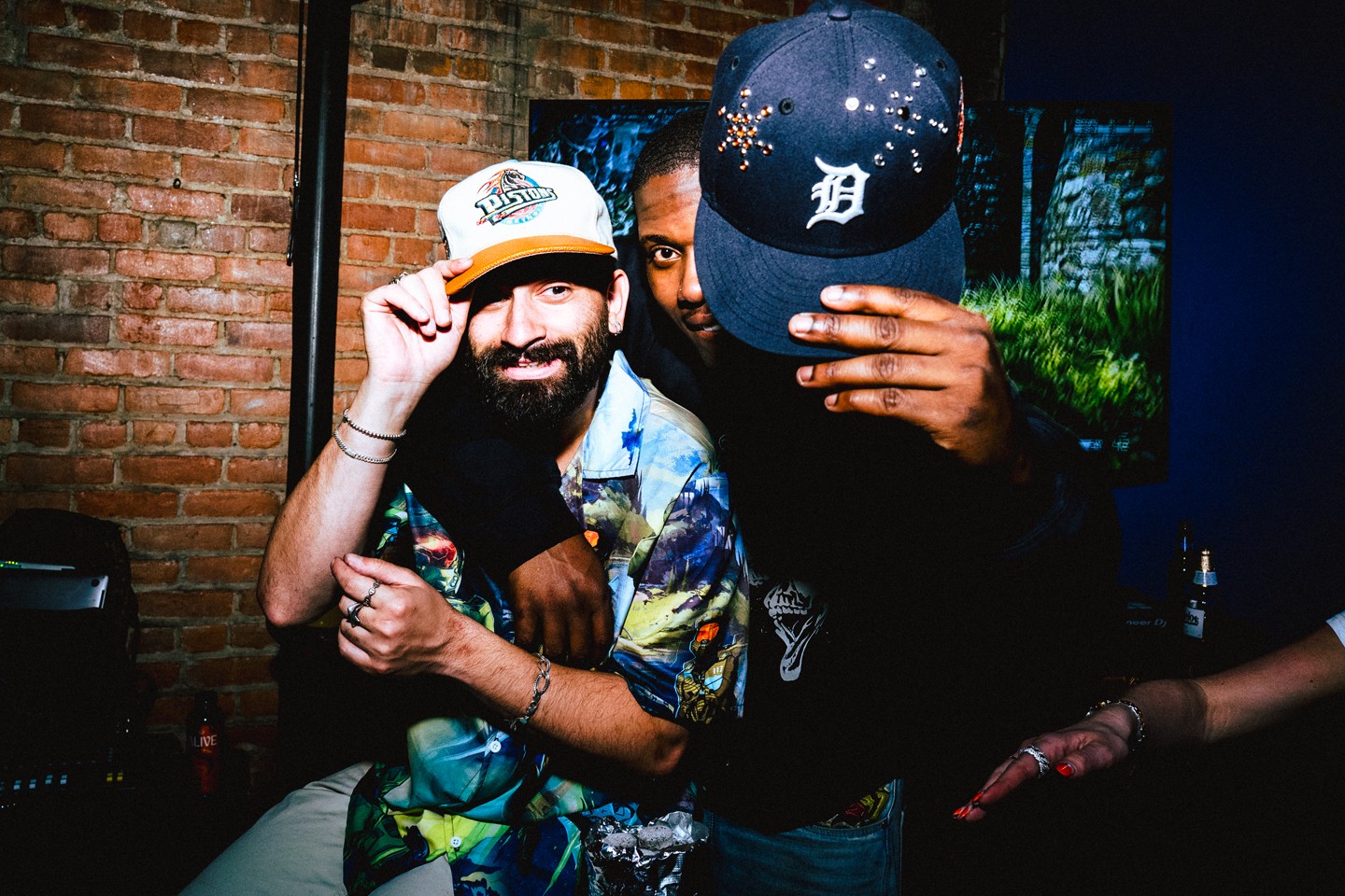 Three people in a dimly lit room with a brick wall, two wearing caps, one with a beard, smiling and posing.
