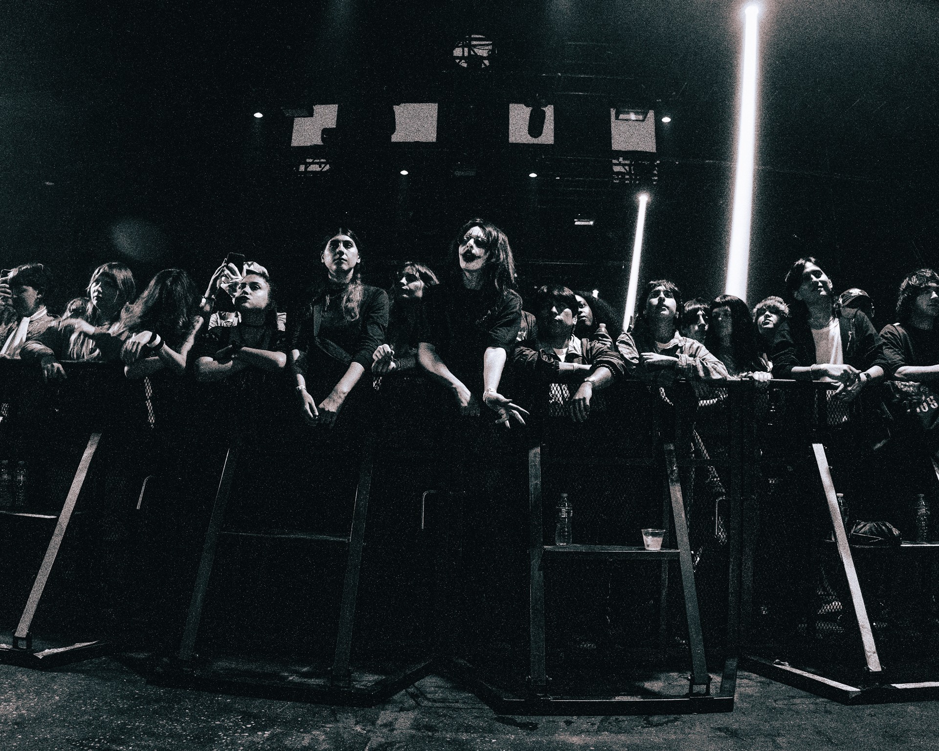Group of people standing behind a barrier in a dark setting, illuminated by a vertical light source, with stage lights above.