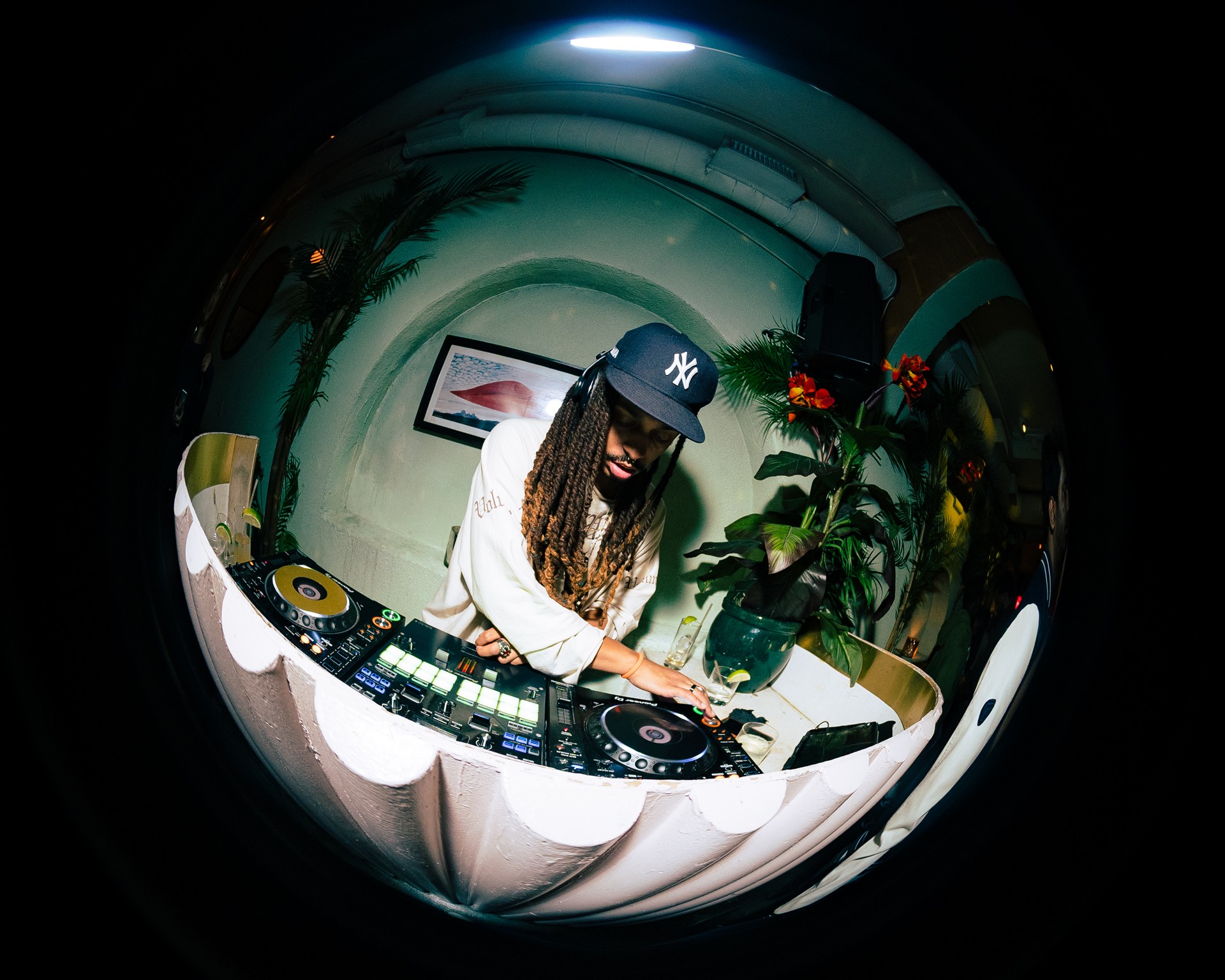 Person wearing a baseball cap DJing with equipment and a plant in the background.