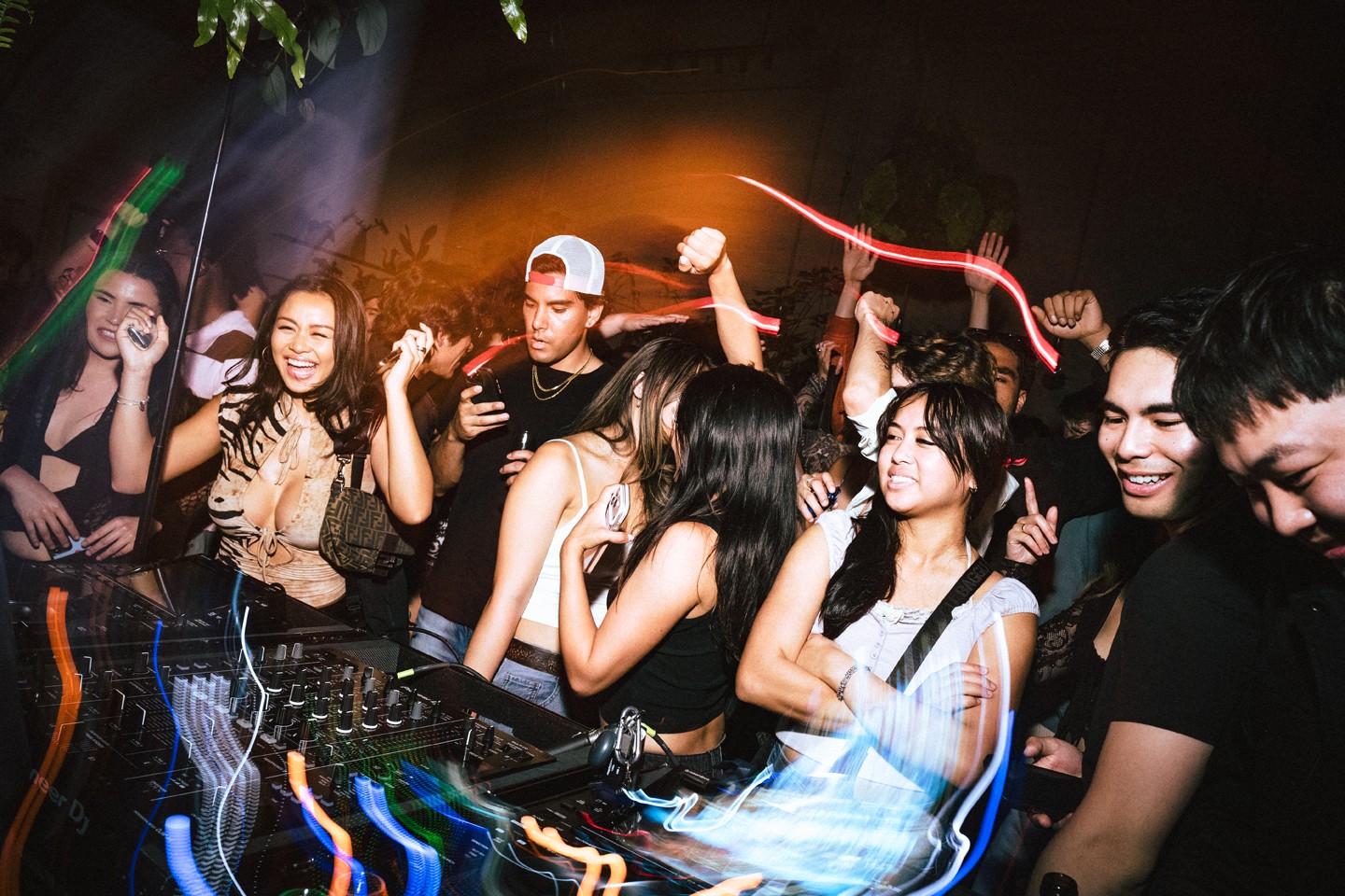 Group of young people dancing and enjoying music at a party or club, with colorful lights and a DJ booth.
