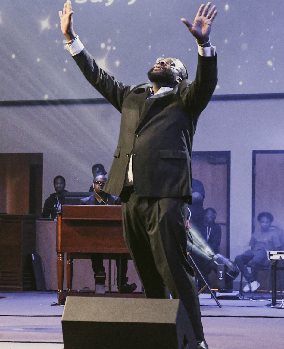 Man in suit with arms raised on stage, audience in background, stage lights shining down.