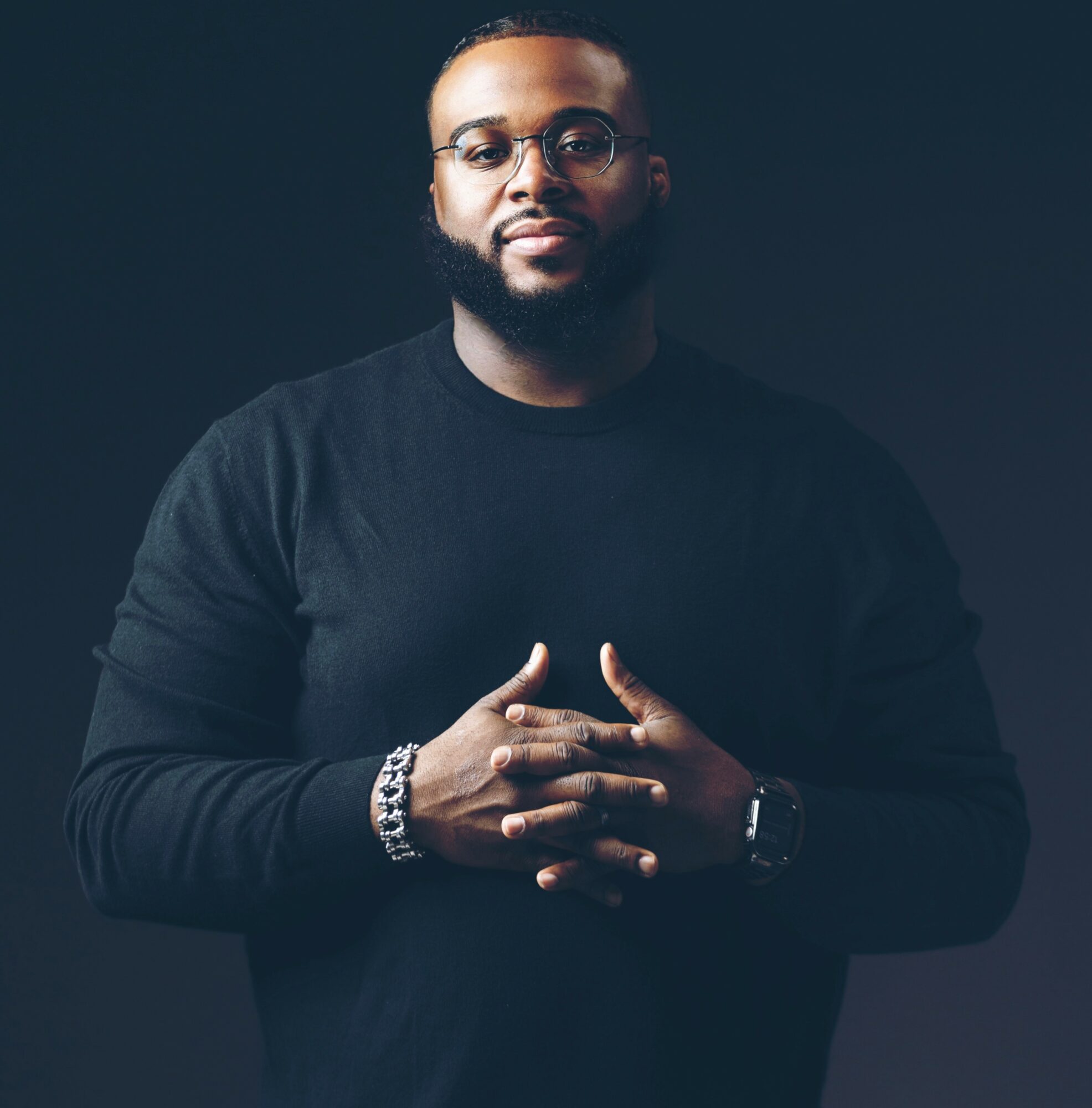 Man with glasses and beard wearing a black shirt, hands clasped, dark background