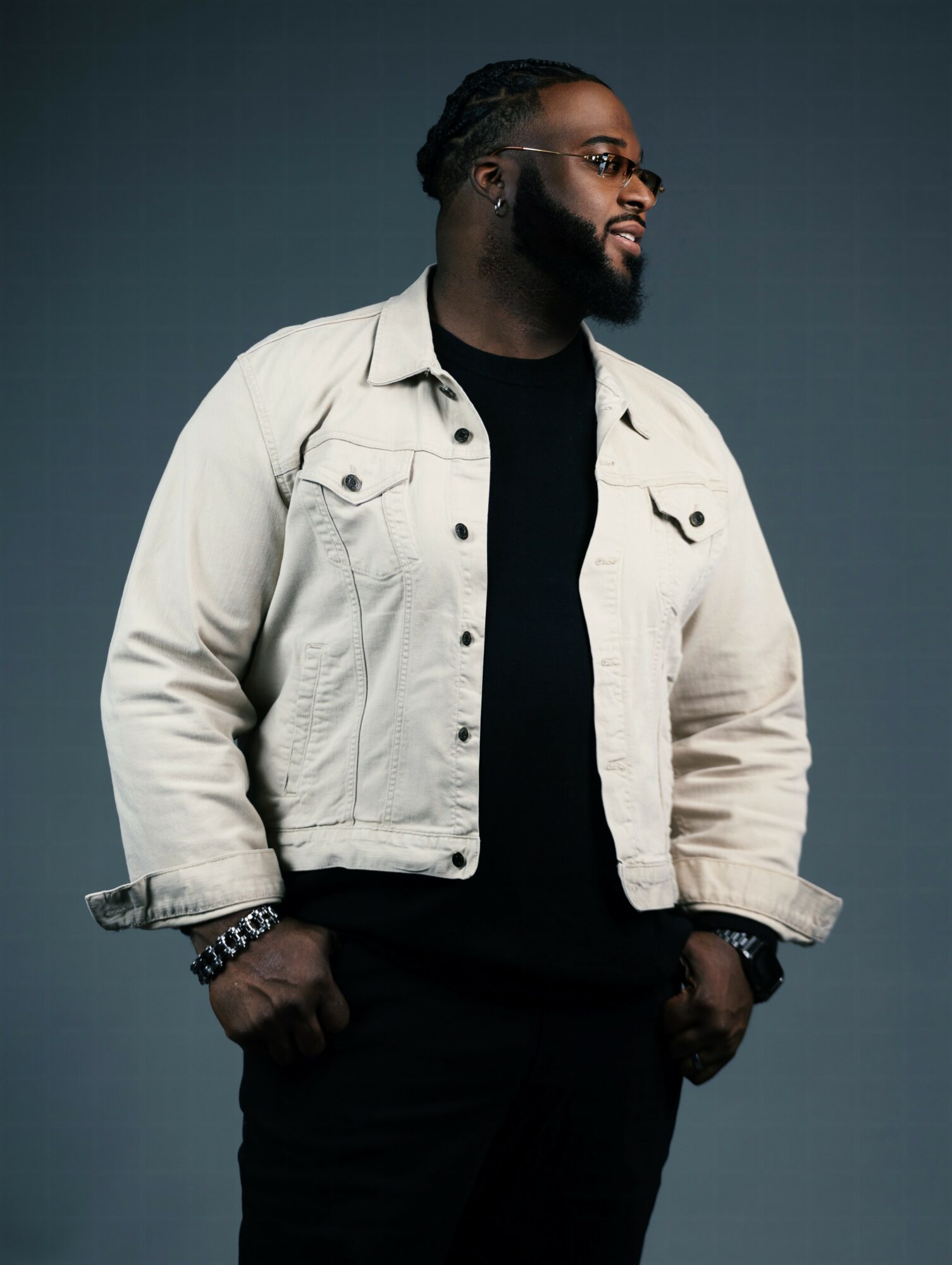 Man with glasses and beard wearing a light jacket over a black shirt, standing against a dark background.