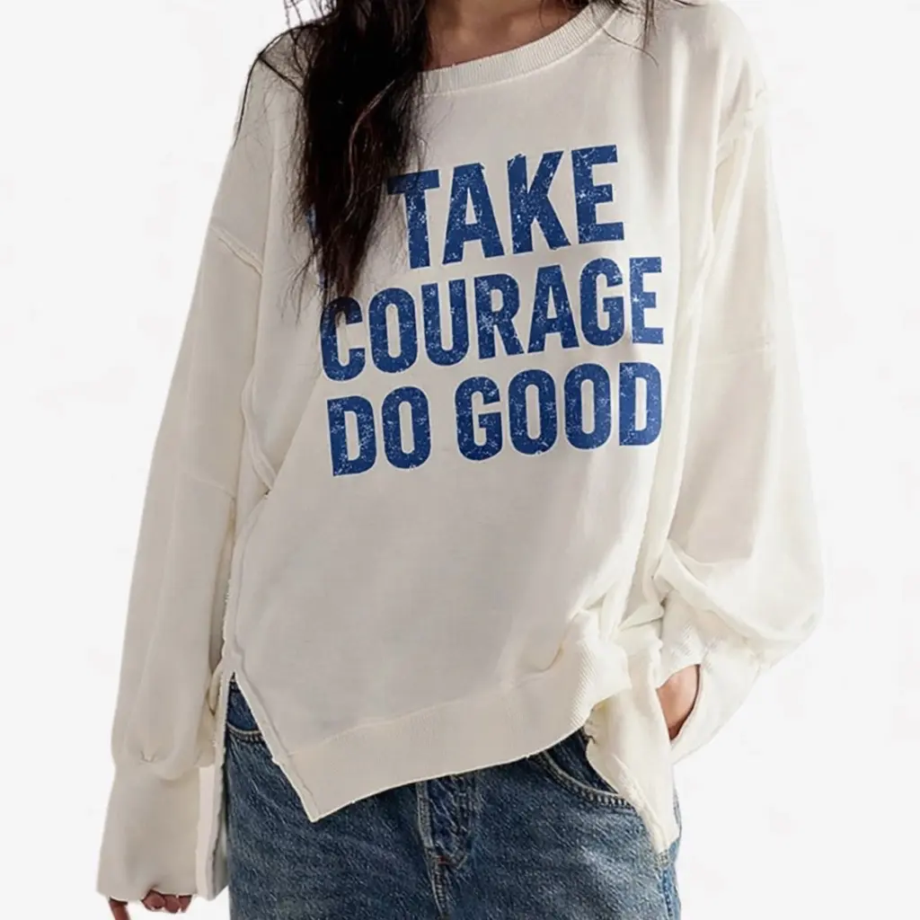 Person wearing a white sweatshirt with blue text, standing with one hand in pocket, against a plain background.