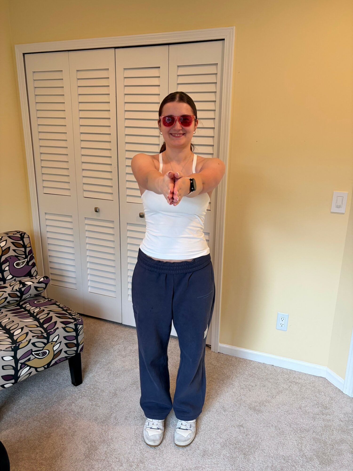 Person taking a mirror selfie, wearing sunglasses, white tank top, dark pants, and sneakers, standing in a room with yellow walls and a closed closet door.