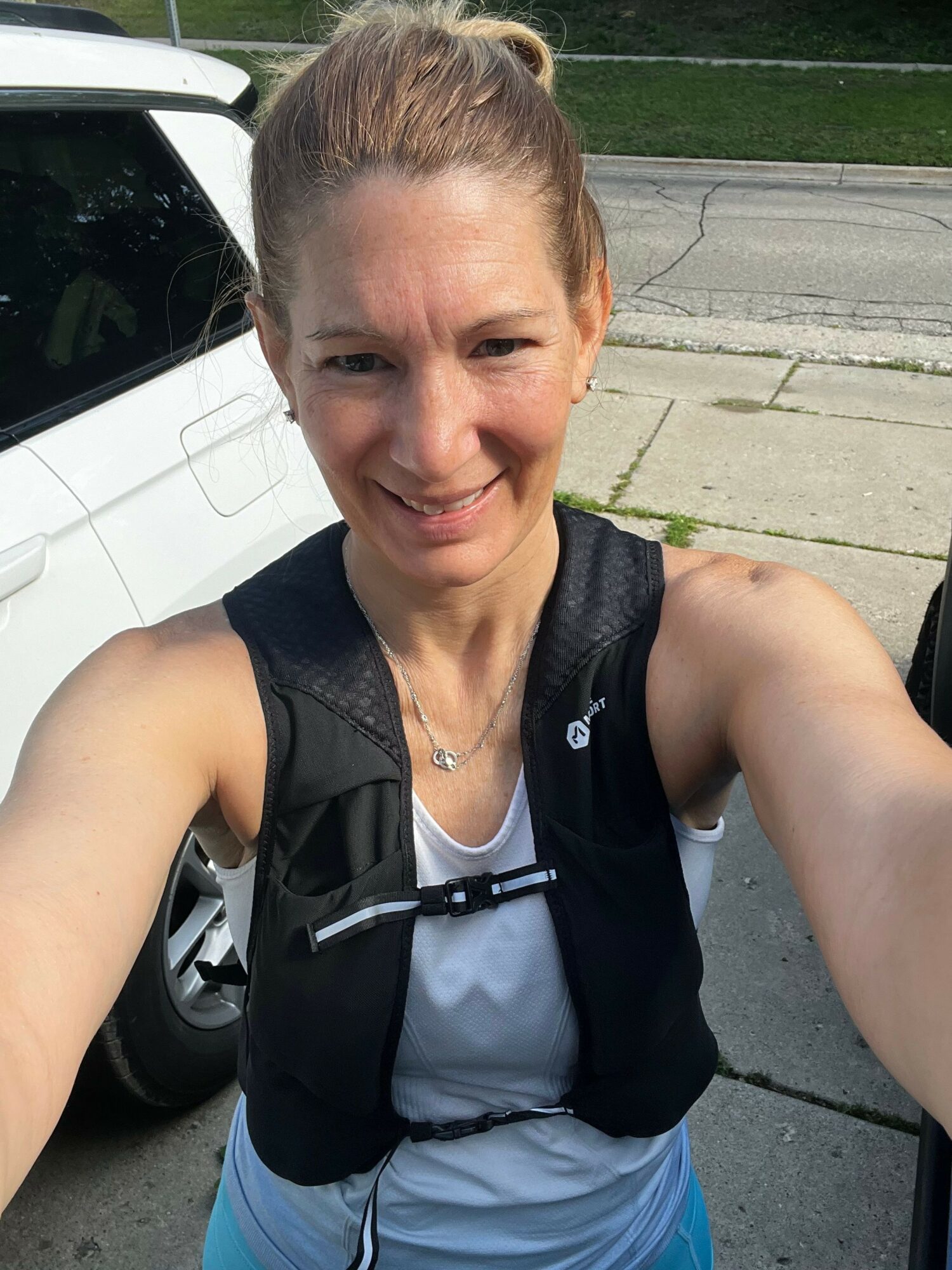 Woman with blonde hair tied back, smiling, wearing a black vest and light-colored shirt, outdoors on a sidewalk.