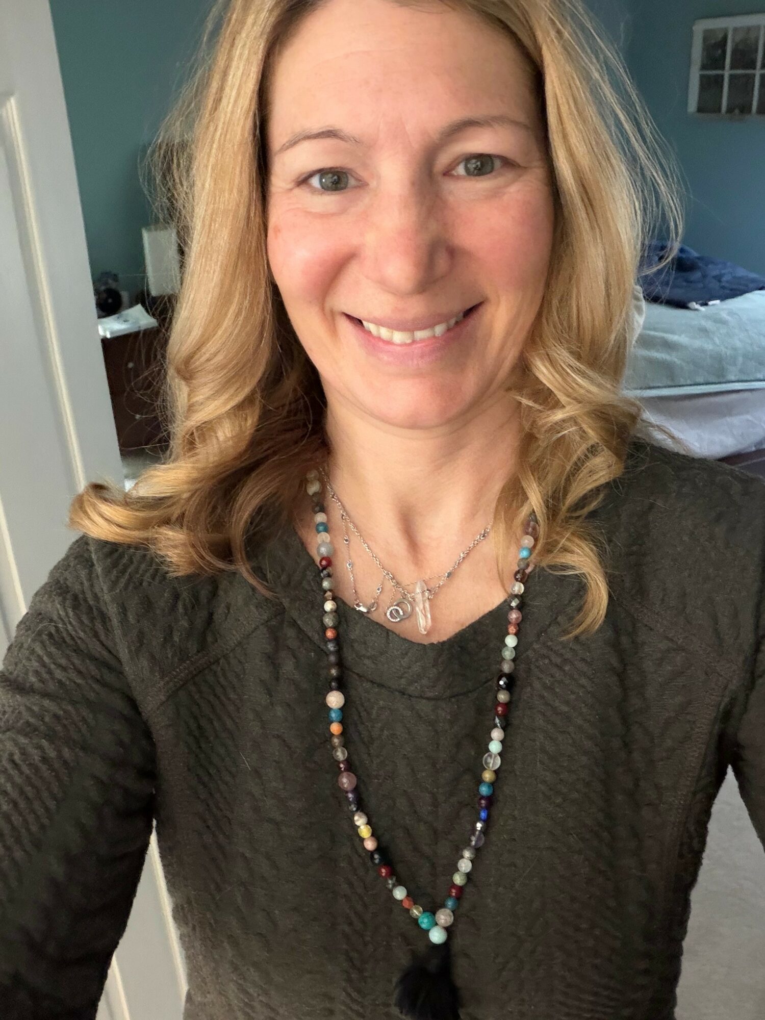 Smiling woman with blonde hair wearing a dark sweater and colorful beaded necklaces, standing in a room with a bed in the background.