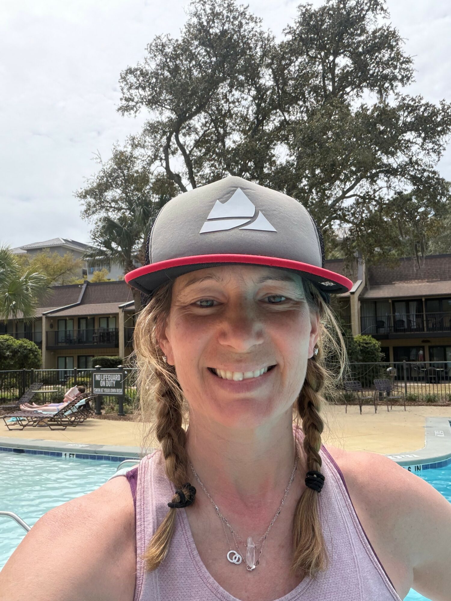 Smiling woman with braided hair wearing a cap near a swimming pool, trees, and buildings in background.