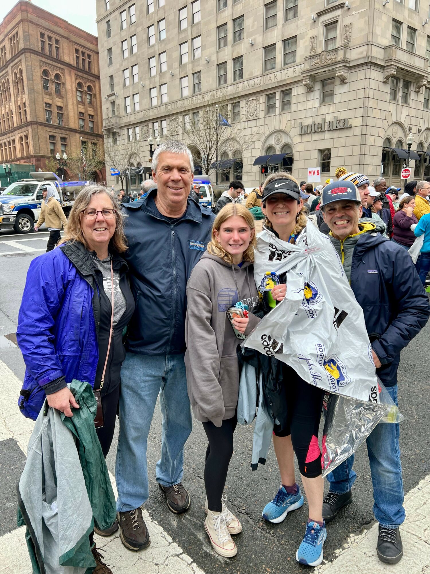 Group of five people standing outdoors in an urban area, some holding flowers and a large wrapped gift, with buildings and cars in the background.