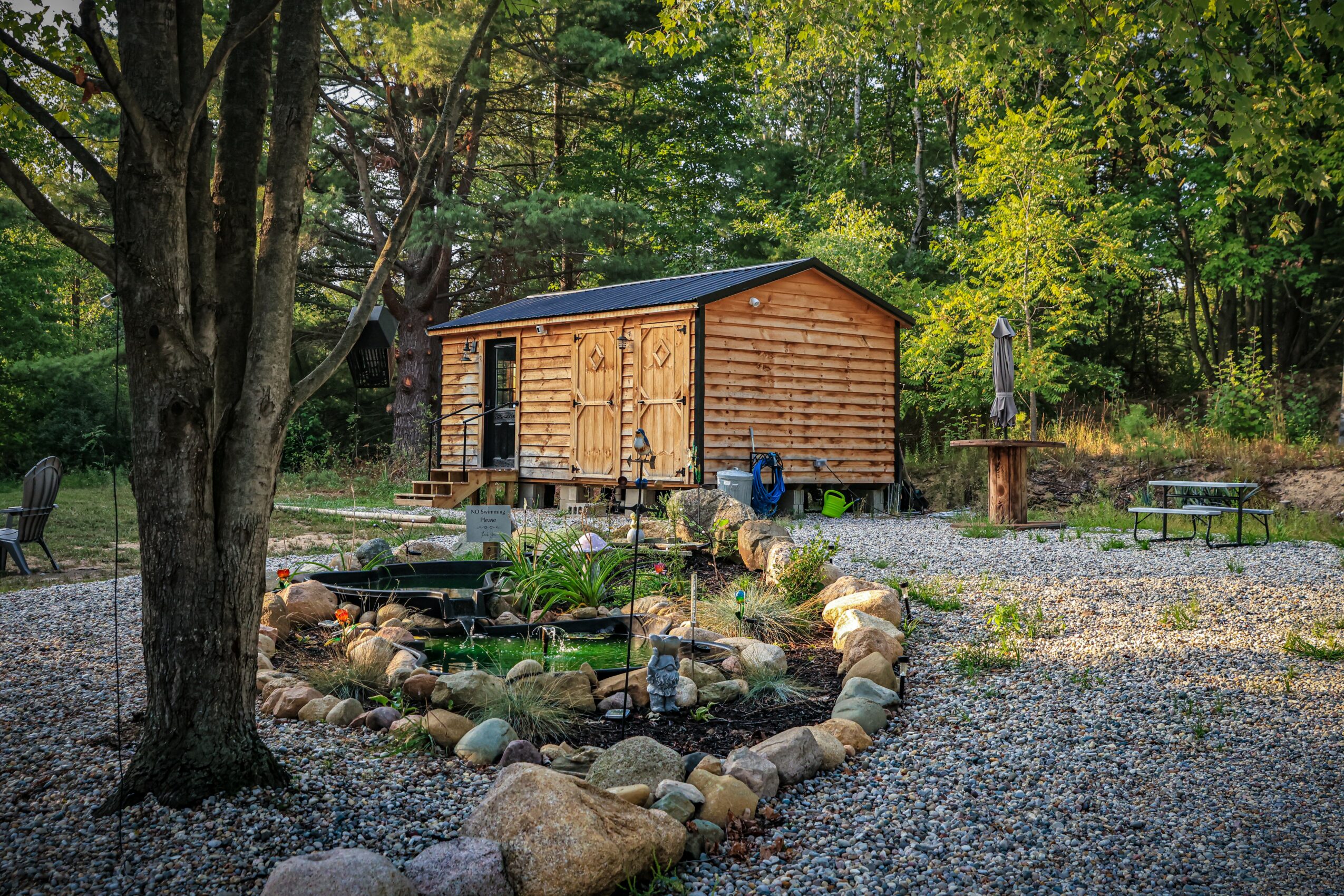 Wooden shed in a garden with trees, a pond surrounded by rocks, and outdoor seating areas.