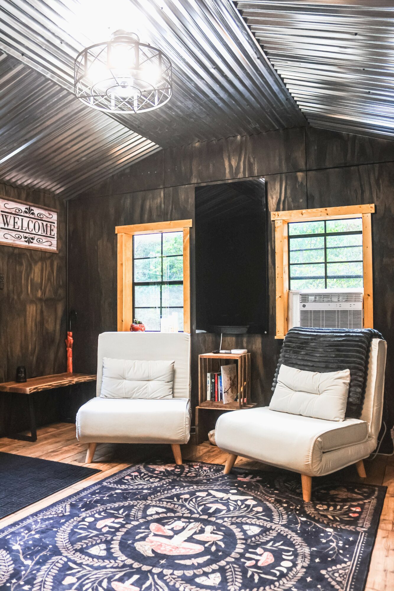 Interior room with two chairs, windows, bookshelf, and a patterned rug, wooden walls, and a ceiling light fixture.