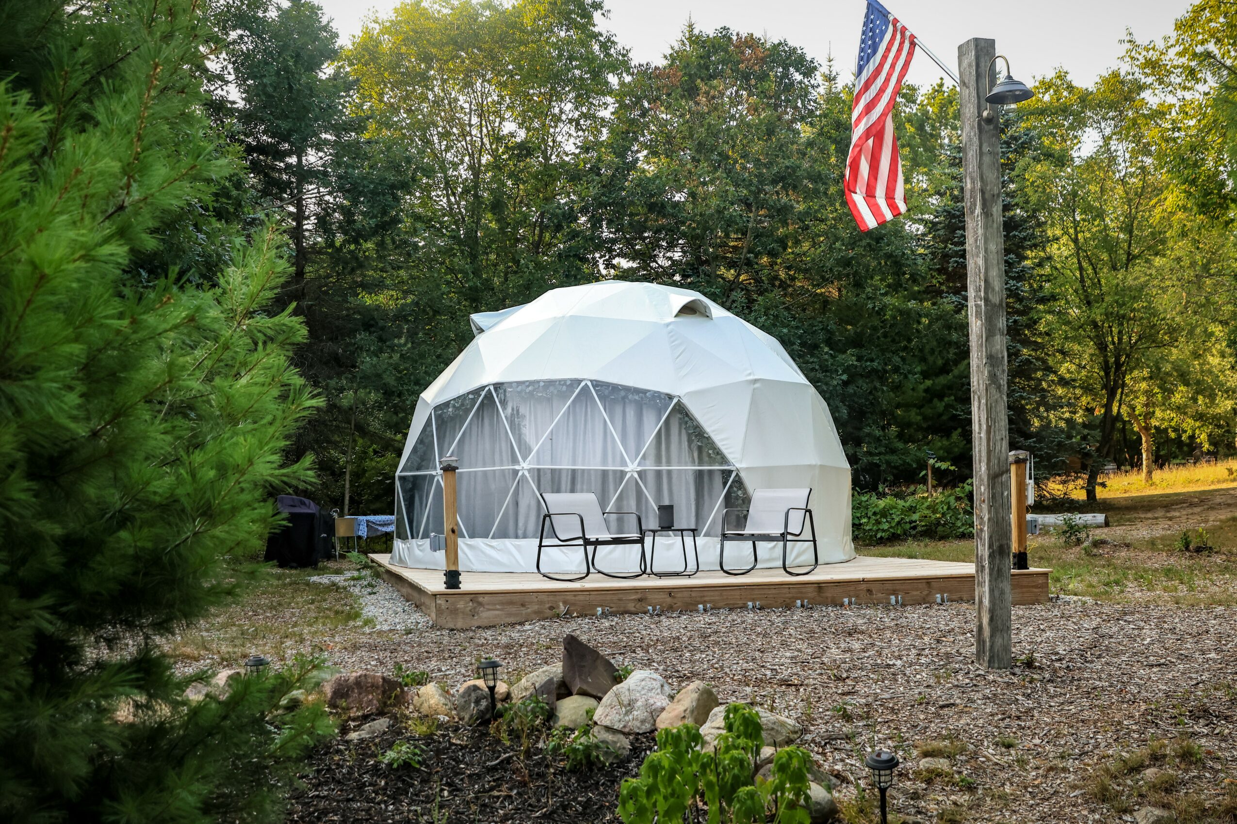 Geodesic dome with chairs on a wooden deck, American flag on pole, trees in background, outdoor setting.