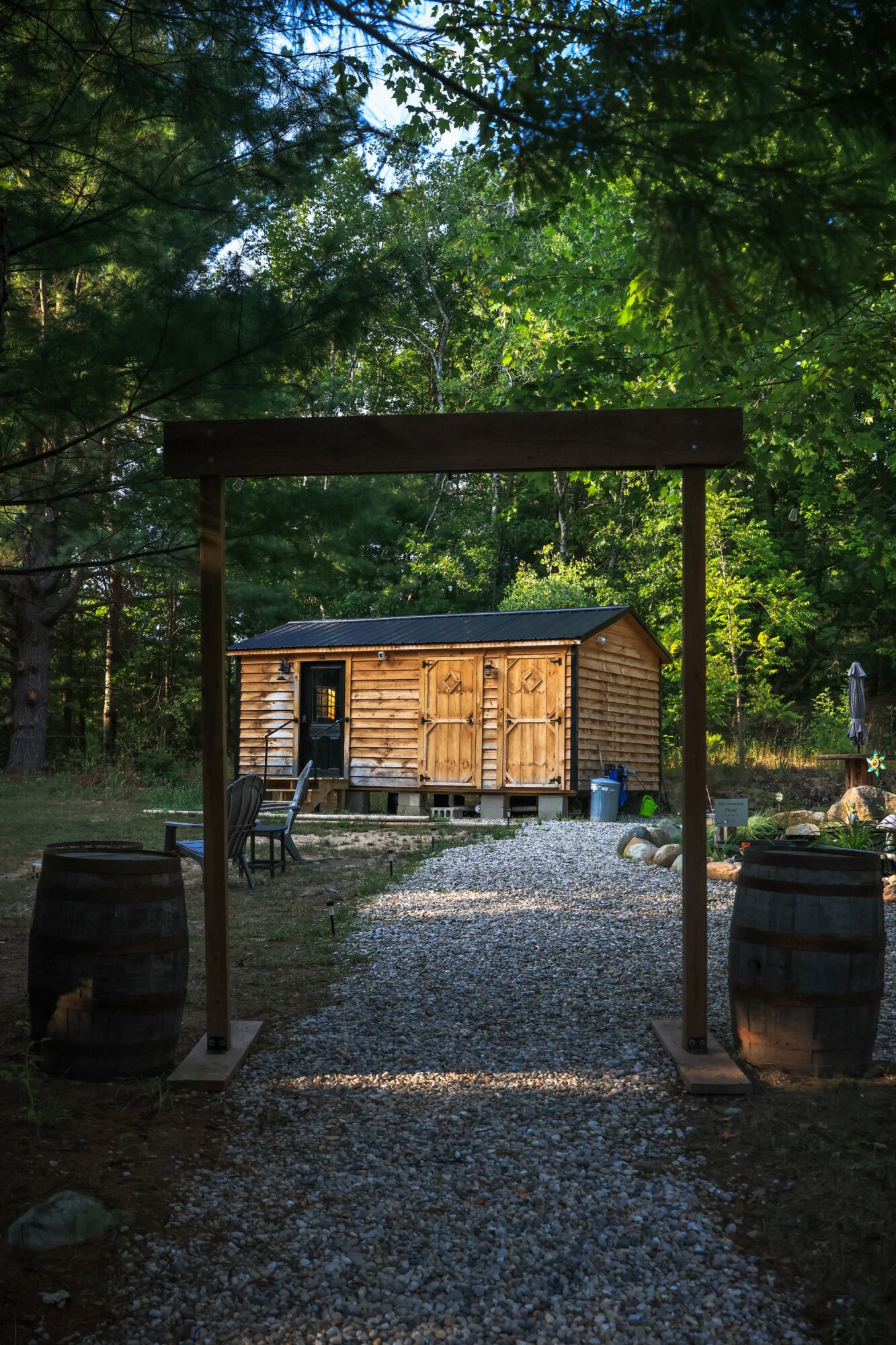 Wooden shed with a black roof, surrounded by trees and greenery, viewed through a wooden archway with barrels on sides.