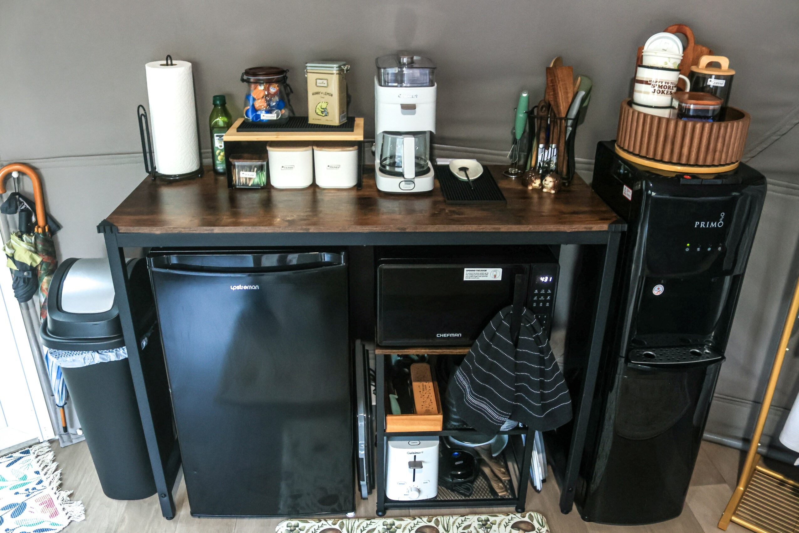 Kitchen counter with coffee maker, cups, and utensils, black mini fridge, microwave, and water cooler, against a gray wall.