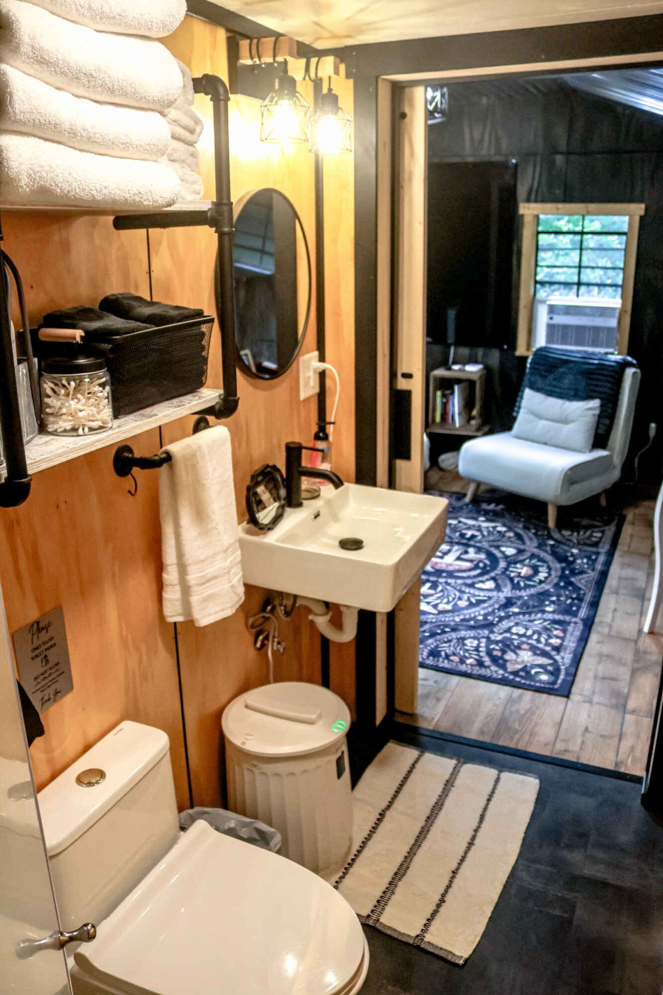 Bathroom with sink, mirror, towels, and toilet, leading to a room with a chair and window, wood flooring, and a rug.