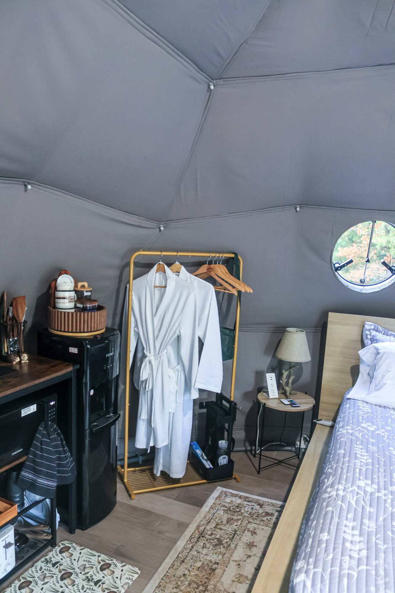 Bedroom with a bed, nightstand, lamp, and clothing rack inside a tent-like structure with a small round window.