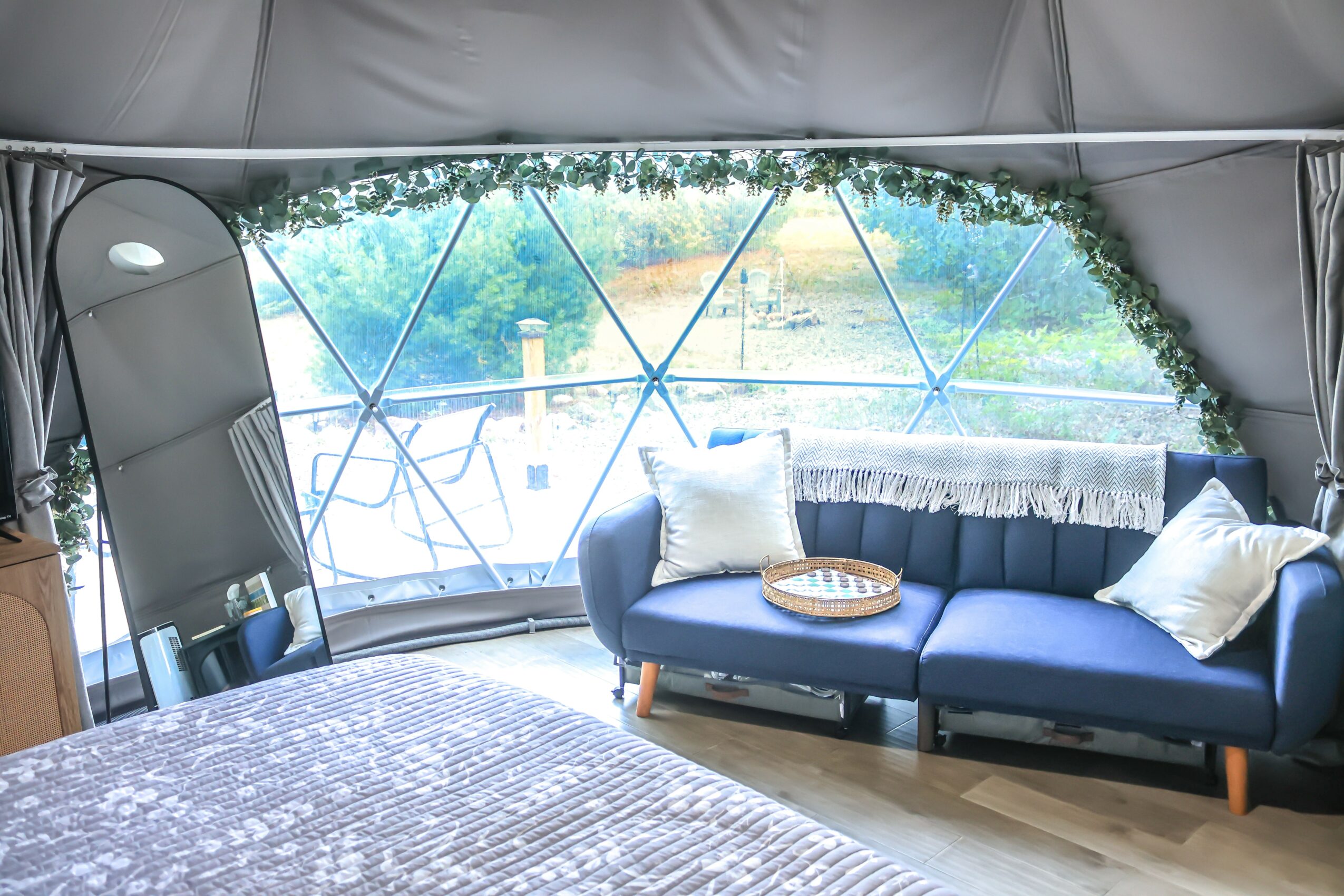 Interior of a tent with a large window, blue sofa with pillows, and a view of greenery outside.
