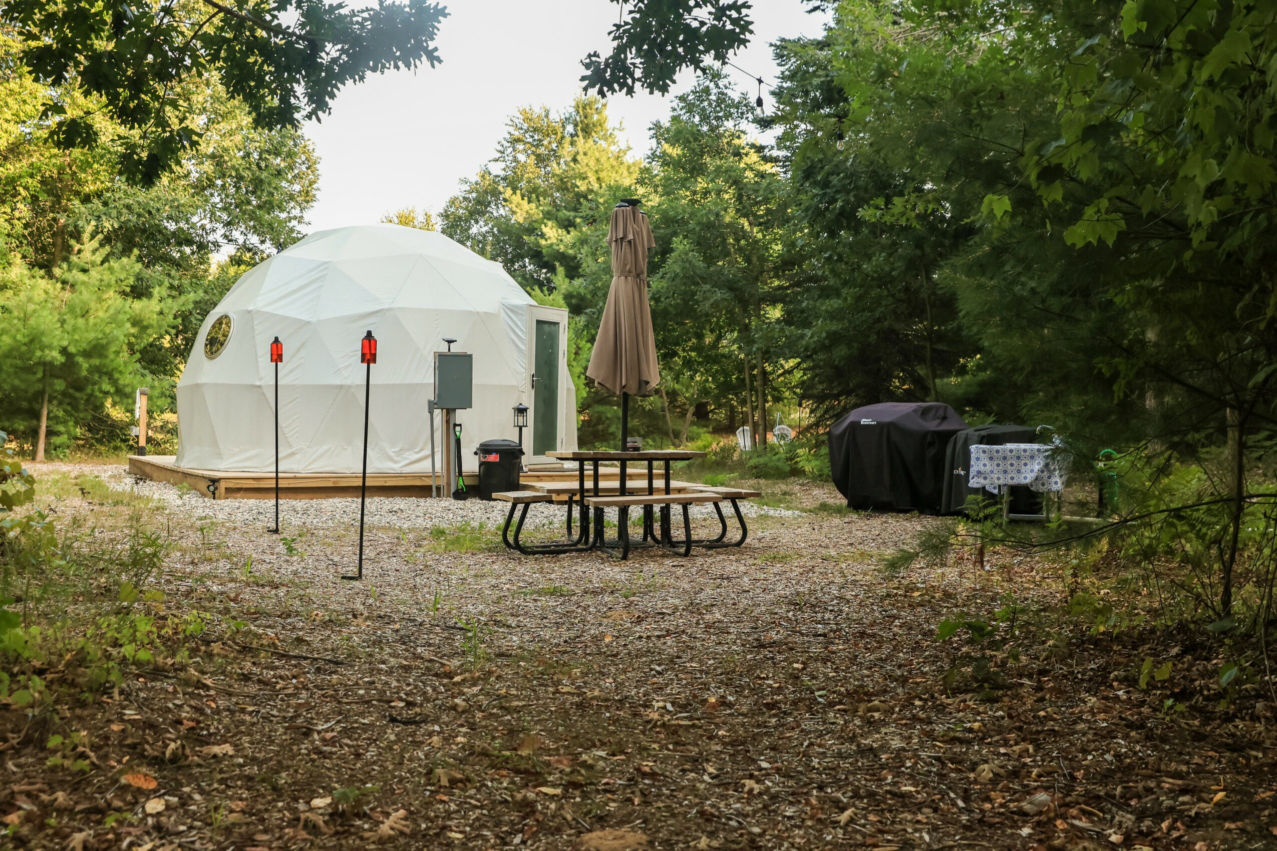Camping area with a white dome tent, picnic table, umbrella, and grill surrounded by trees and greenery.