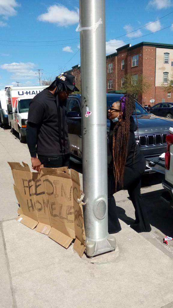Two people standing on sidewalk near a pole, holding a cardboard sign, with parked cars and buildings in background.