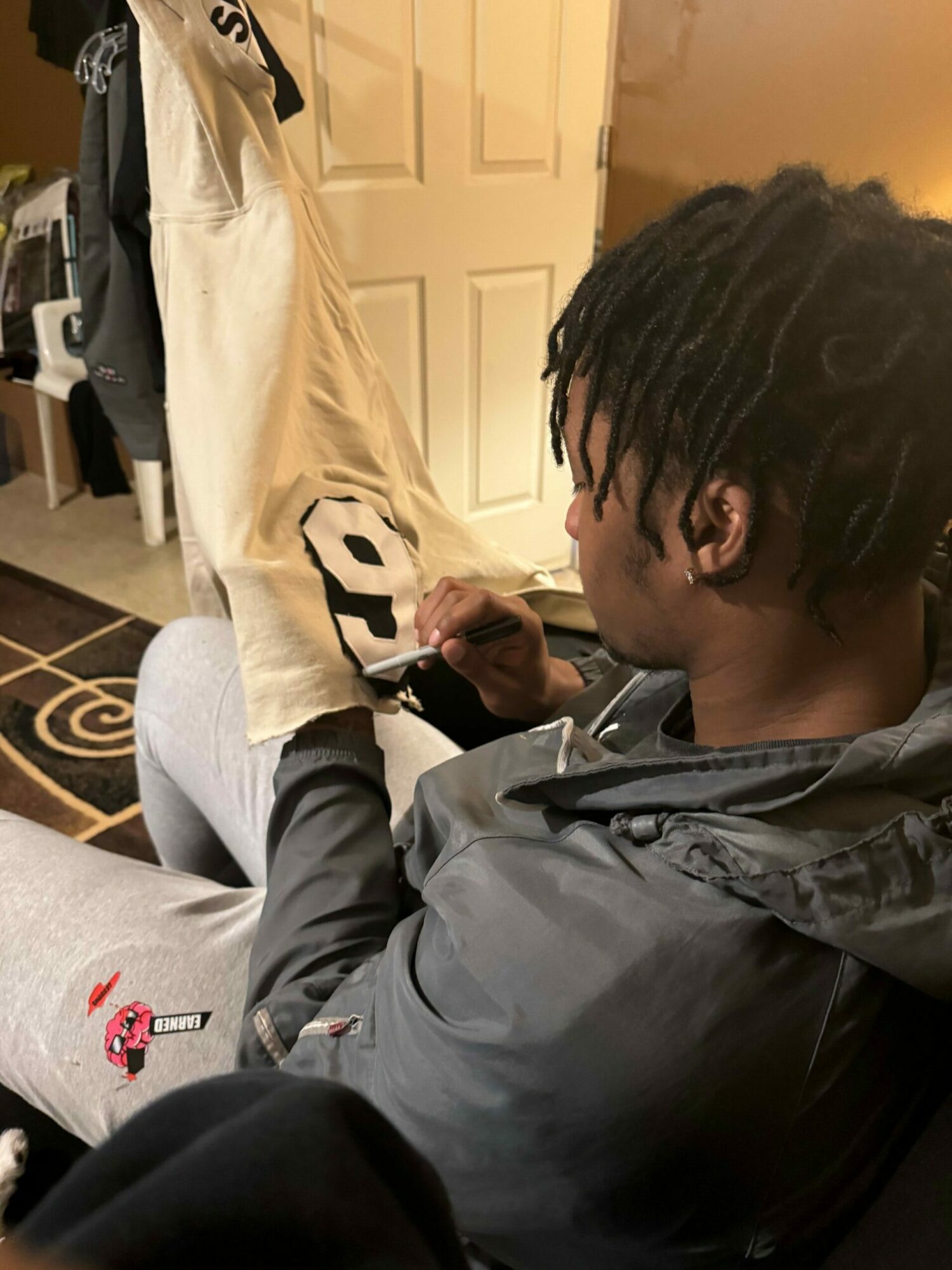 Person with dreadlocks leaning over a table, writing on a piece of paper, in a room with a door and patterned carpet.