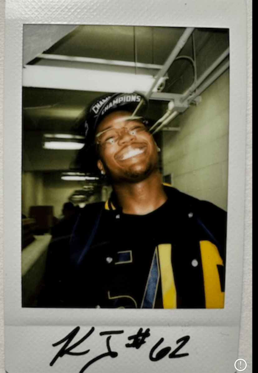Smiling person wearing a cap and jacket in an indoor setting, with a hallway background.