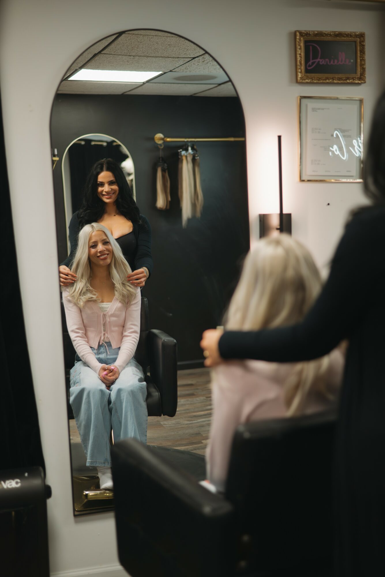 Two women in a hair salon, one sitting and one standing, with a mirror reflecting their images, and framed pictures on the wall.