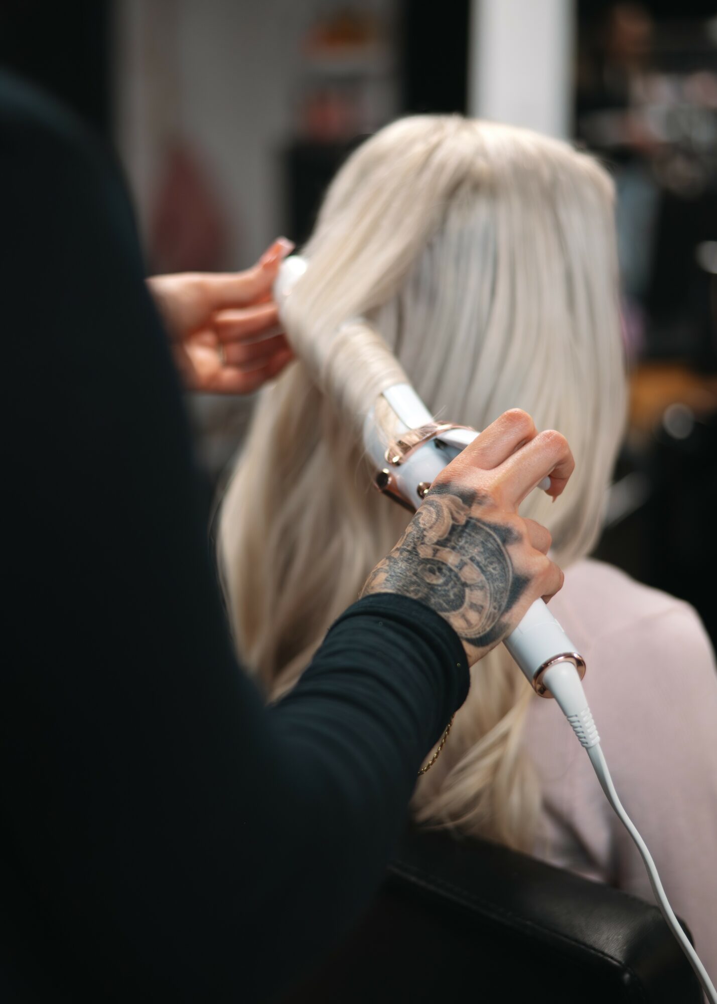 Person with tattooed arm styles long, platinum blonde hair in a salon chair, using a curling iron on the hair.