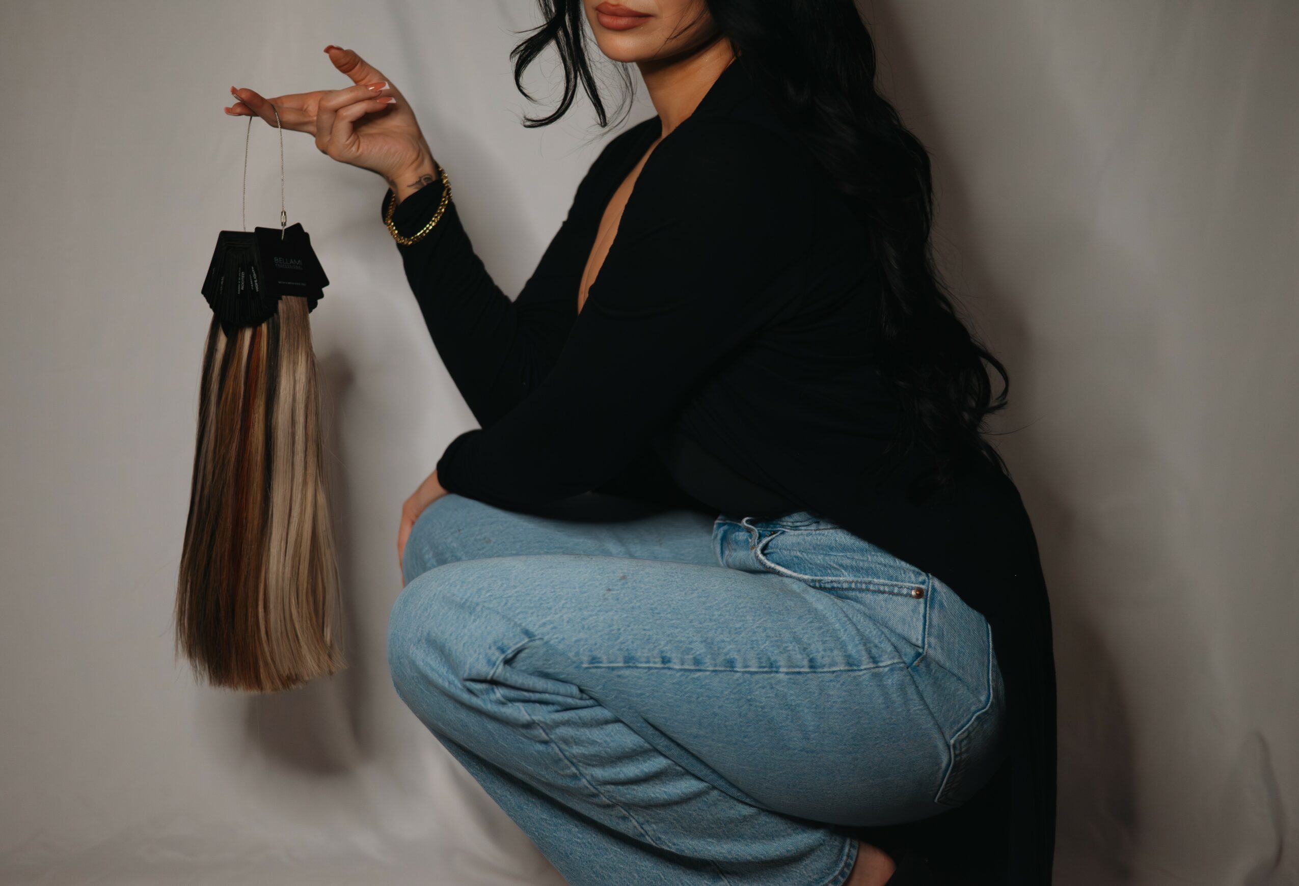 Person crouching with long hair, holding a hair extension bundle, wearing a black top and light blue jeans.