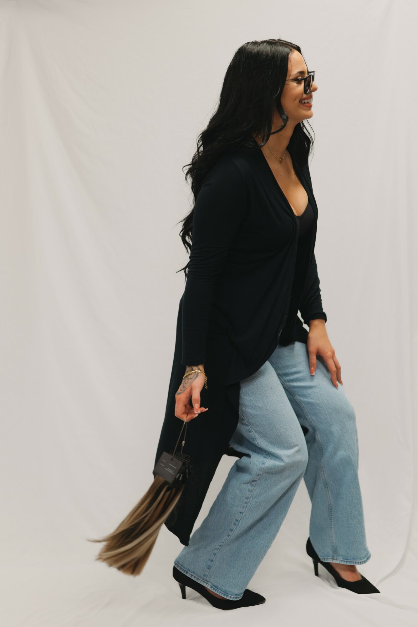 Woman with long dark hair wearing glasses, black top, light blue jeans, and black high heels, holding a handbag, standing against a plain background.