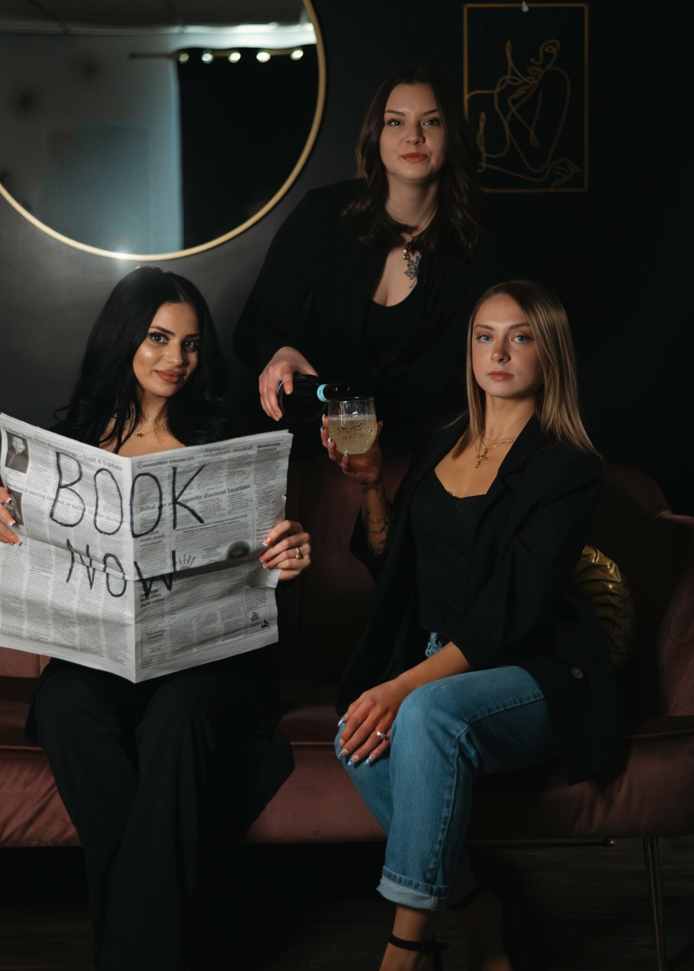 Three women in black clothing in a dimly lit room, one holding a newspaper, another with a drink, and the third standing.