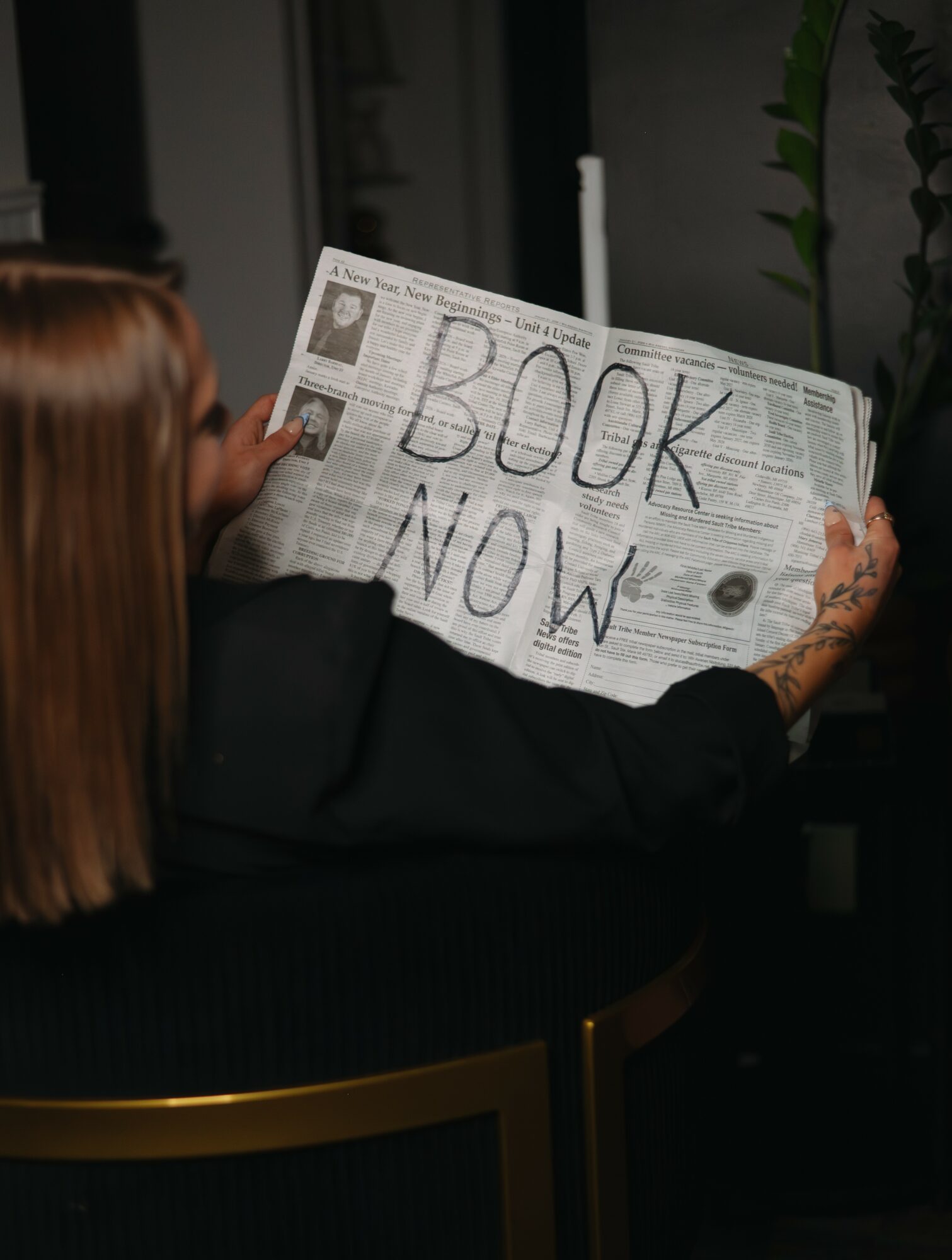 Person with red hair holding a newspaper with large handwritten words 'BOOK NOW' in a room with plants.